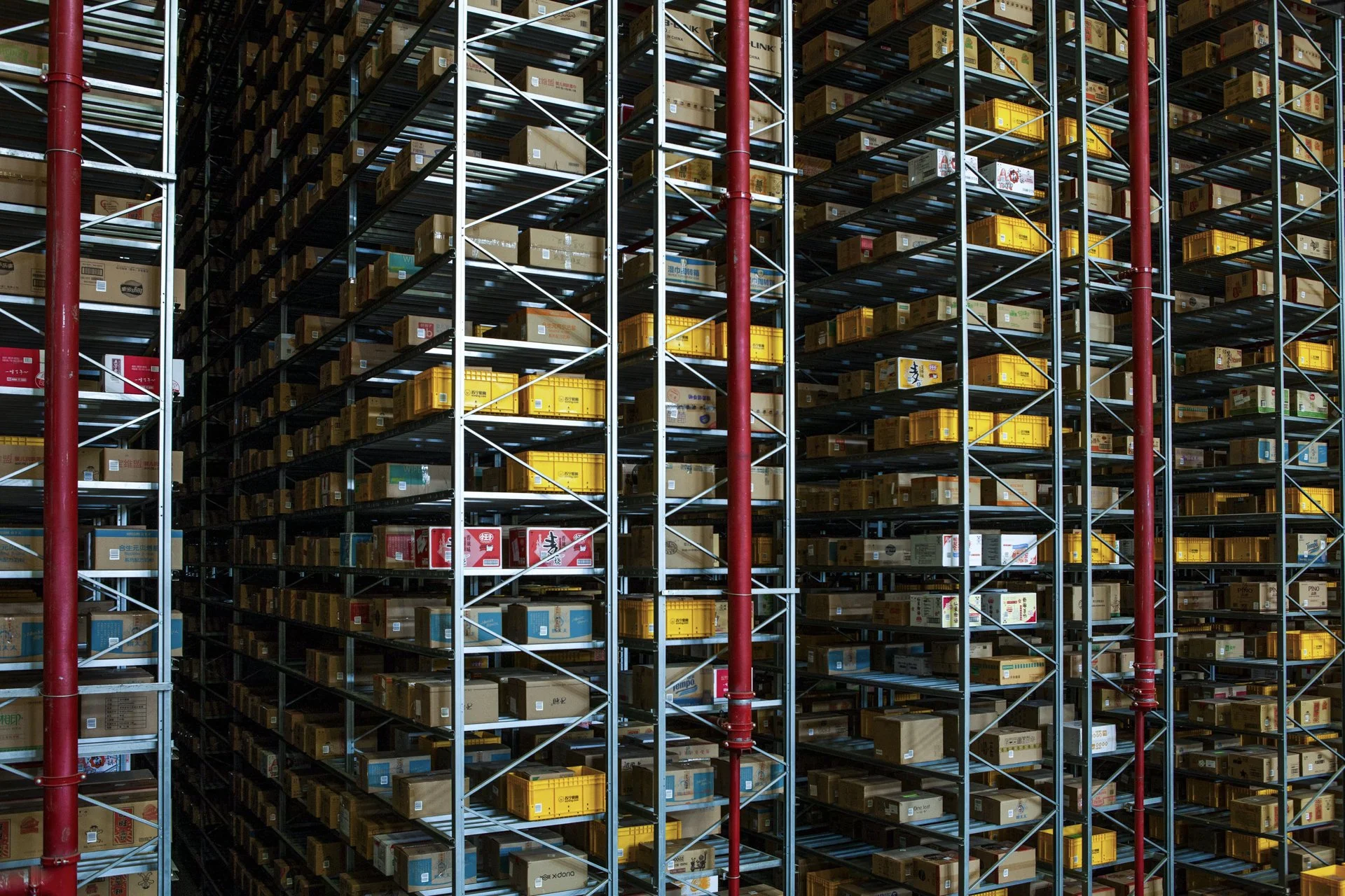 A large warehouse with multiple levels of metal shelves holding various boxes. The structure has red vertical support poles and horizontal beams, with some boxes stacked in yellow and brown cartons.