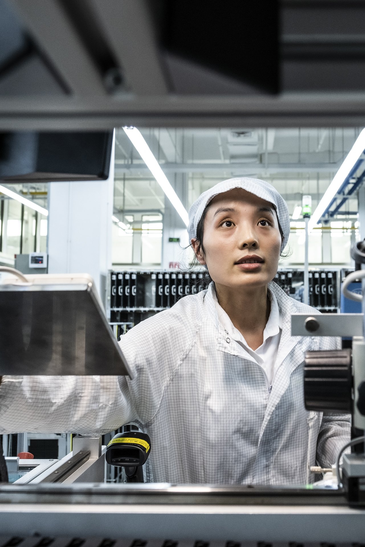 A woman working in a laboratory surrounded by equipment, wearing a white lab coat and a hair cap, with laboratory shelves and lighting in the background.