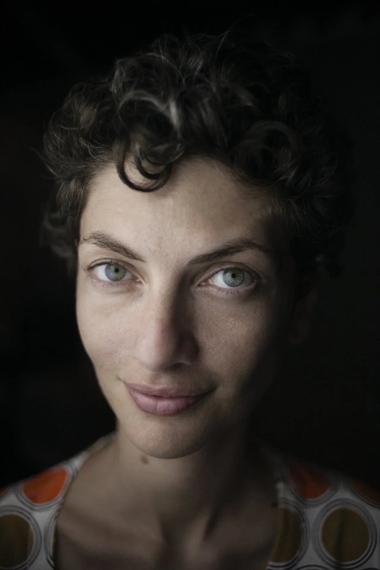 Close-up portrait of a young woman with curly brown hair and blue eyes, smiling gently against a black background, wearing a top with a circular pattern.