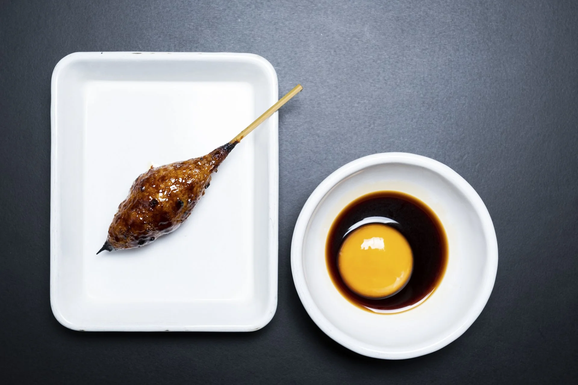 A white rectangular plate with a grilled chicken skewer glazed in sauce, next to a white bowl containing an egg yolk surrounded by soy sauce.