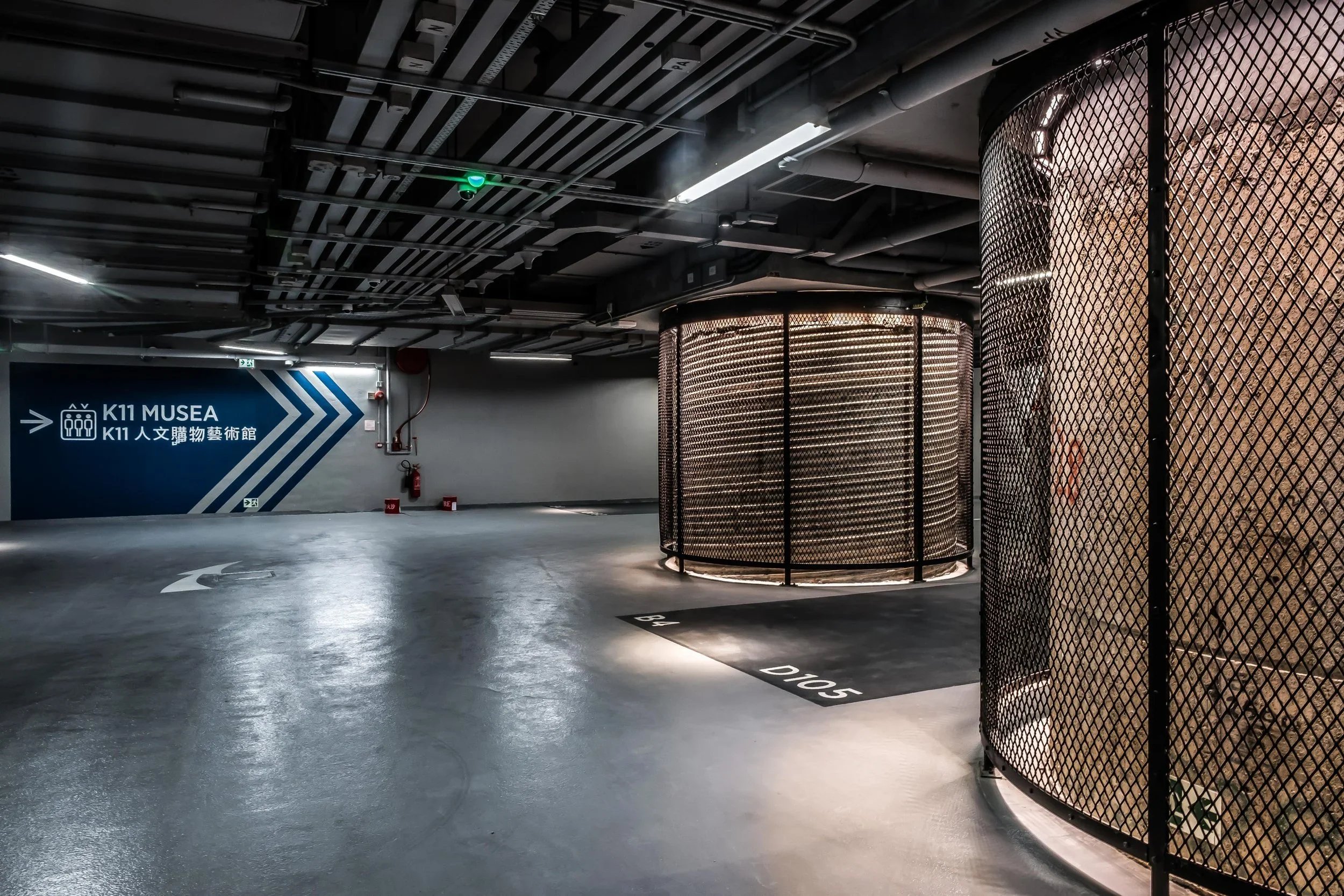 Empty parking garage with a sign pointing to the K11 Musea, showing a directional arrow and Chinese characters. The ceiling has exposed pipes and lighting, with some circular metal structures on the right side.