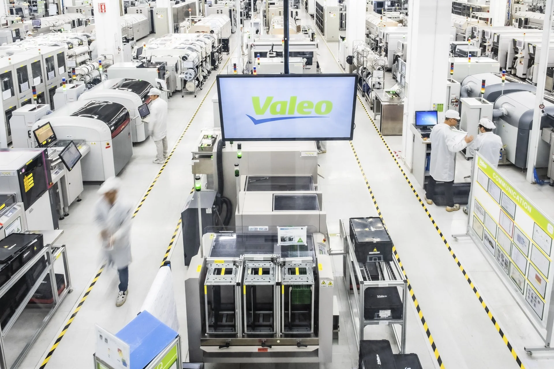 Inside a high-tech electronics manufacturing facility with workers wearing white lab coats and hairnets, operating automated machinery and testing electronic components, with a large Valeo sign on a monitor in the center.