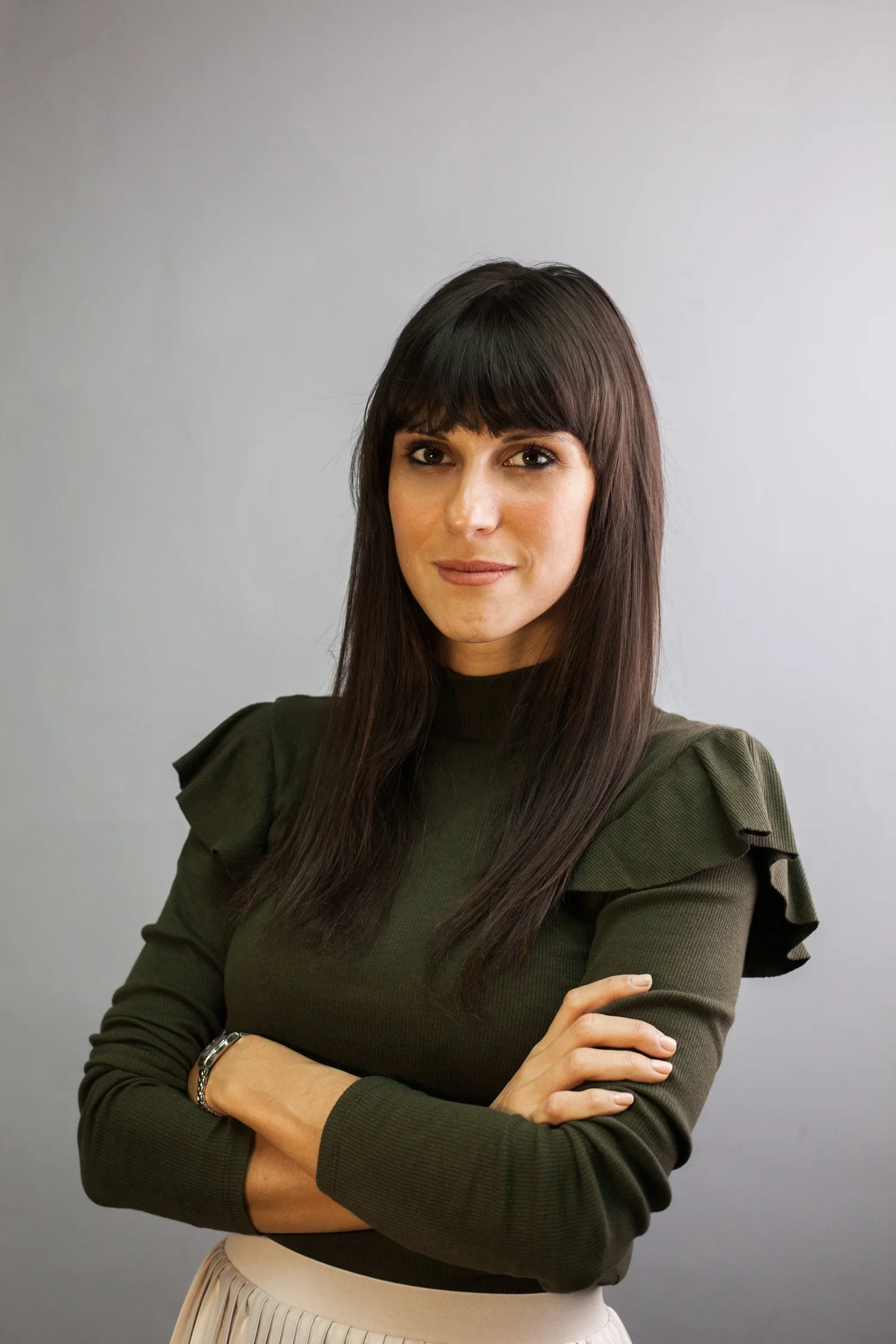 A woman with long dark hair and bangs, wearing a dark olive green top with ruffled sleeves, standing against a plain light gray background. She has her arms crossed and is looking at the camera with a slight smile.