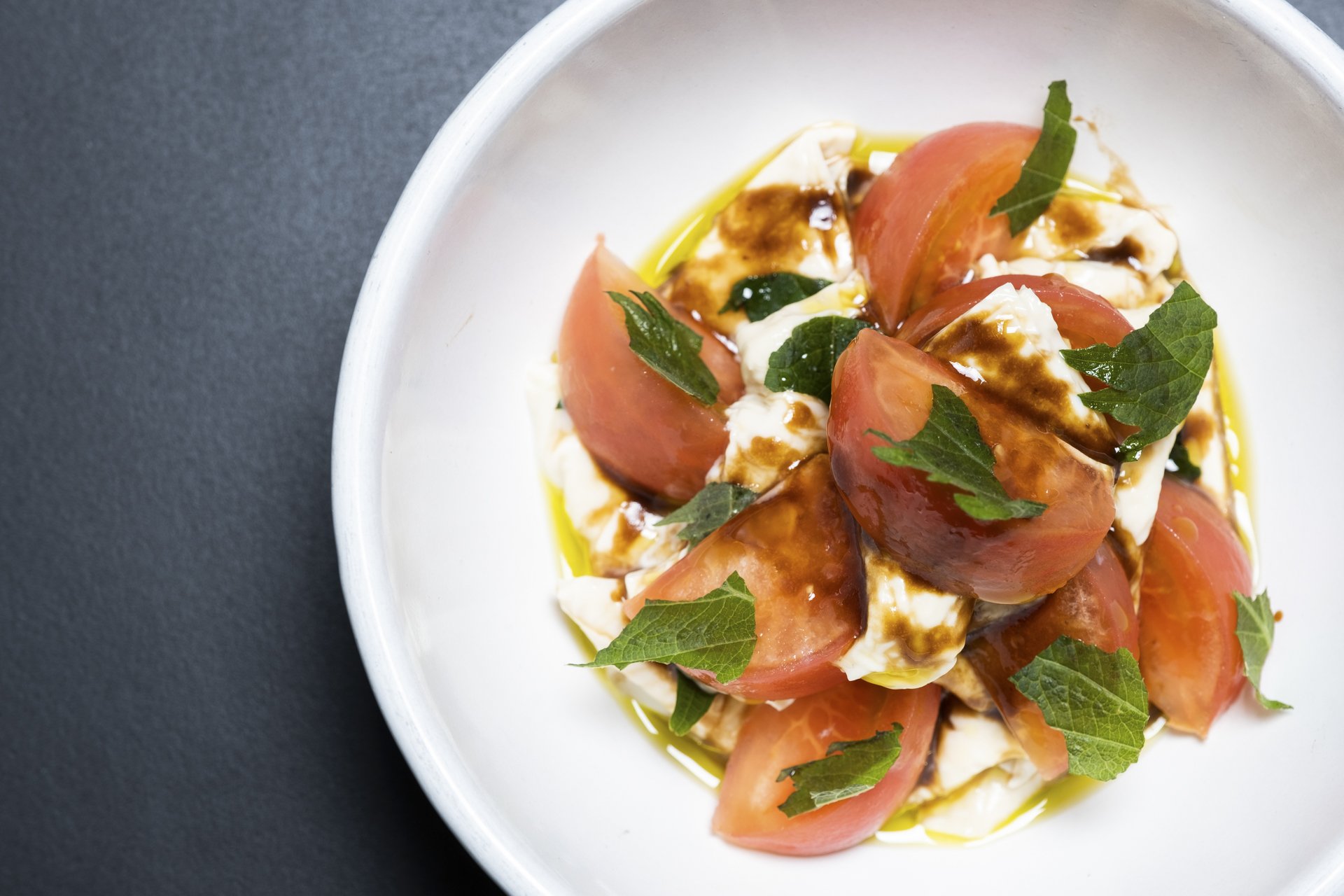 A dish of Caprese salad featuring cherry tomatoes, fresh mozzarella cheese, basil leaves, drizzled with balsamic vinegar and olive oil, served in a white bowl on a dark surface.