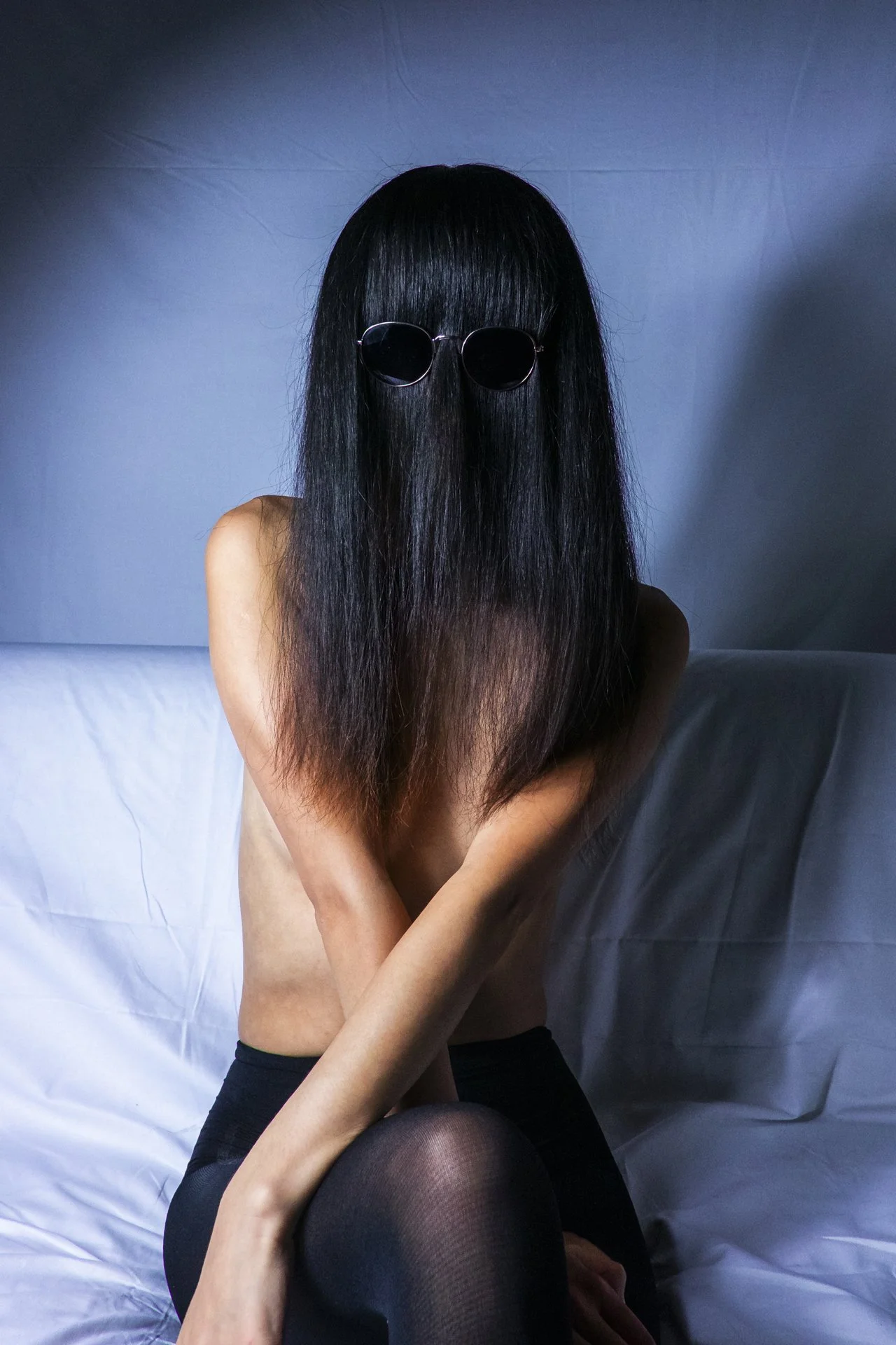 A woman with long black hair wearing sunglasses, sitting on a bed with white sheets, arms crossed, with her face obscured by her hair and sunglasses.