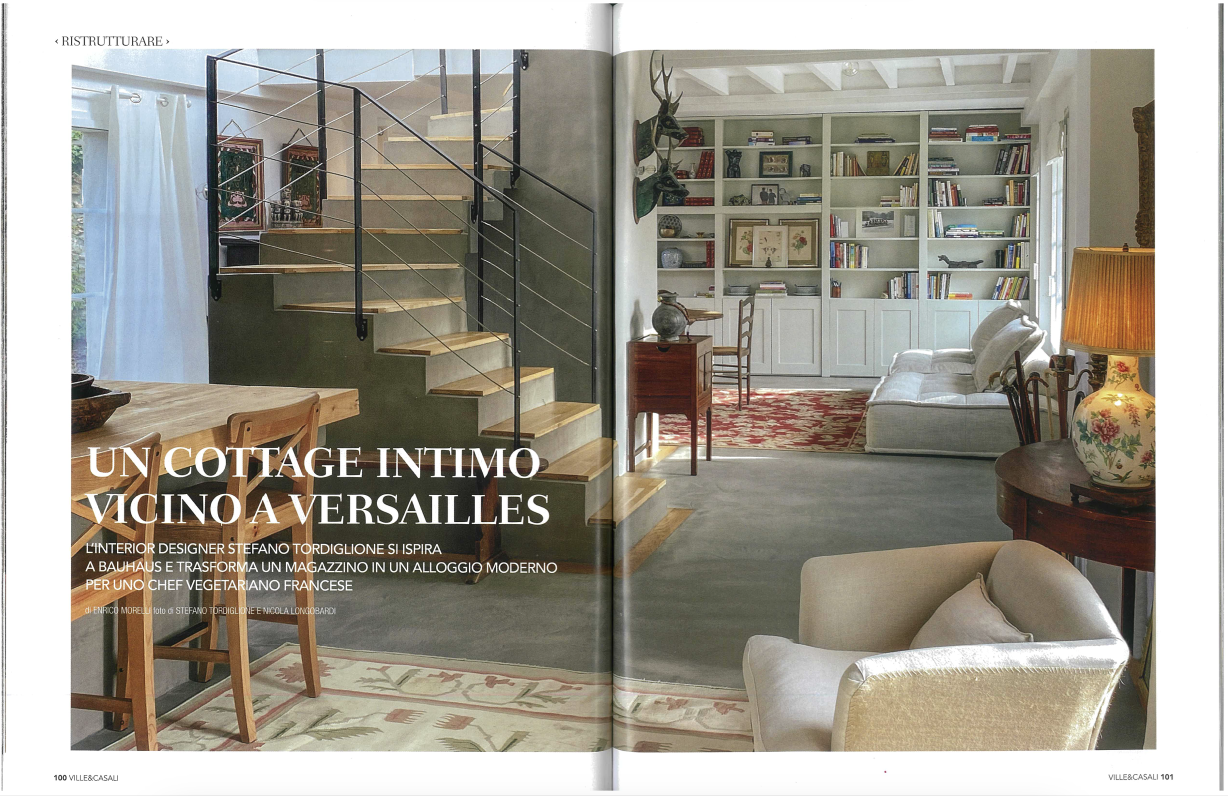 A cozy living room with a staircase on the left, white curtains, a built-in bookshelf on the right filled with books and decorative items, and seating including a beige armchair and a sofa. A lamp with a yellow shade and floral design is on a side ta