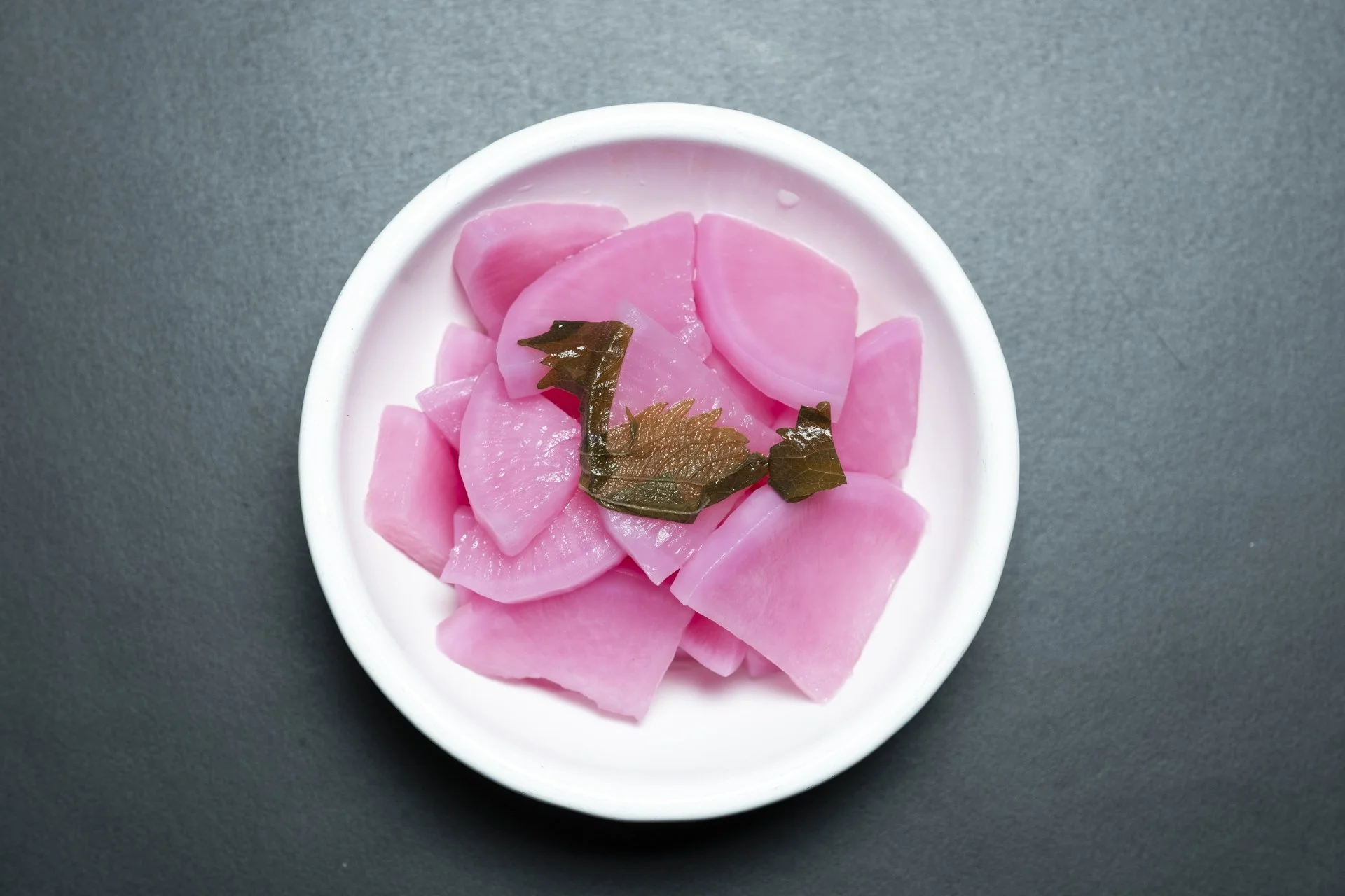 A white bowl containing pickled pink radish slices garnished with green seaweed, placed on a dark surface.