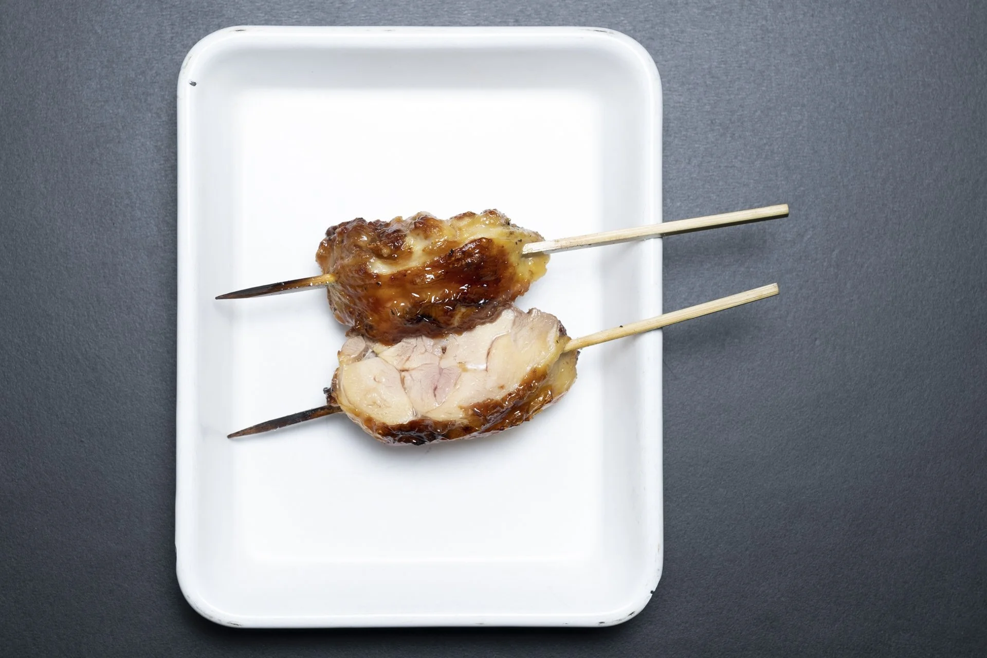 Two skewered pieces of grilled meat on a white rectangular plate, one glazed with sauce, against a dark background.