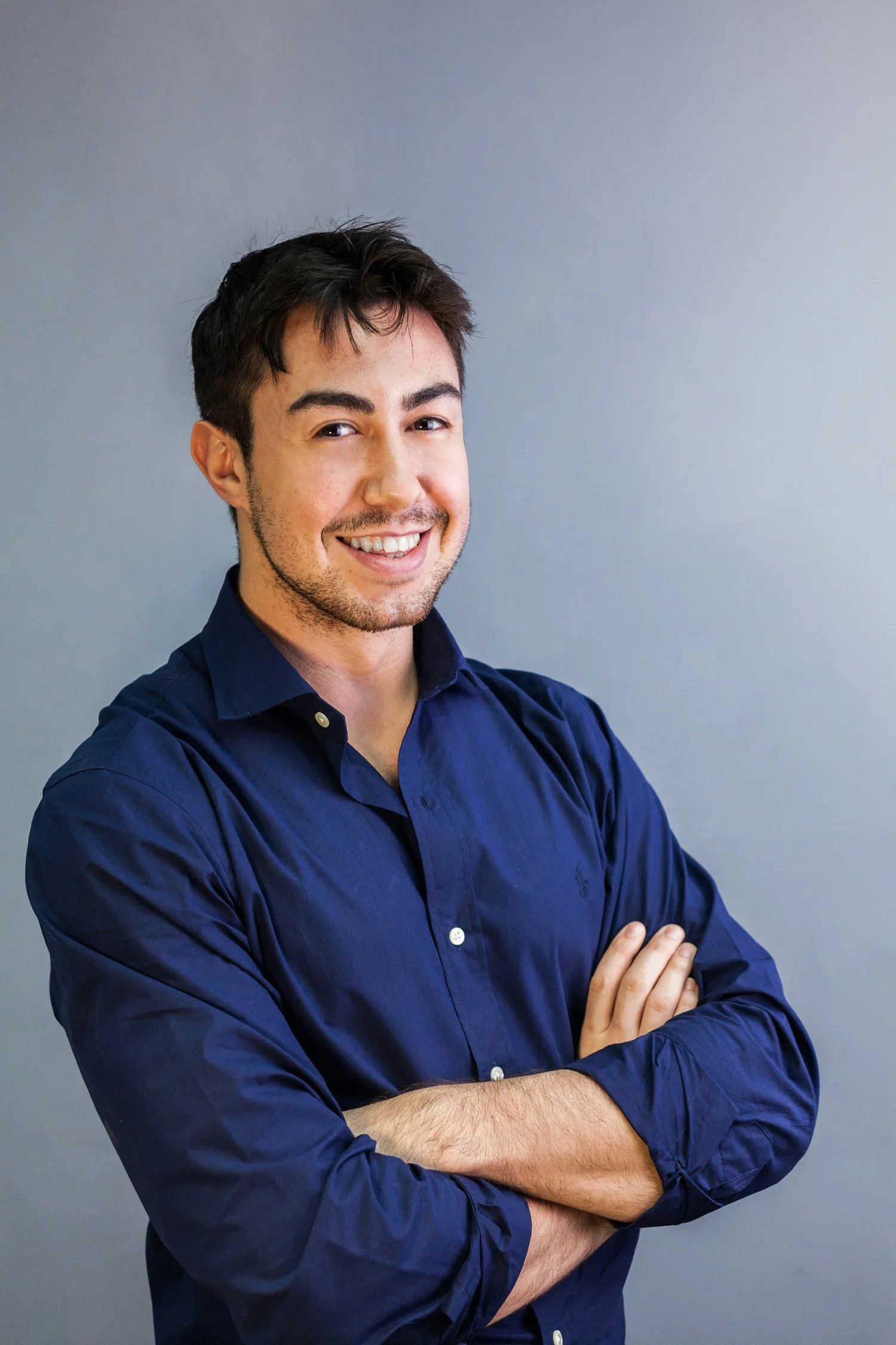 A young man with dark hair, light skin, and a five o'clock shadow, smiling and crossing his arms, wearing a dark blue button-up shirt, standing against a plain gray background.
