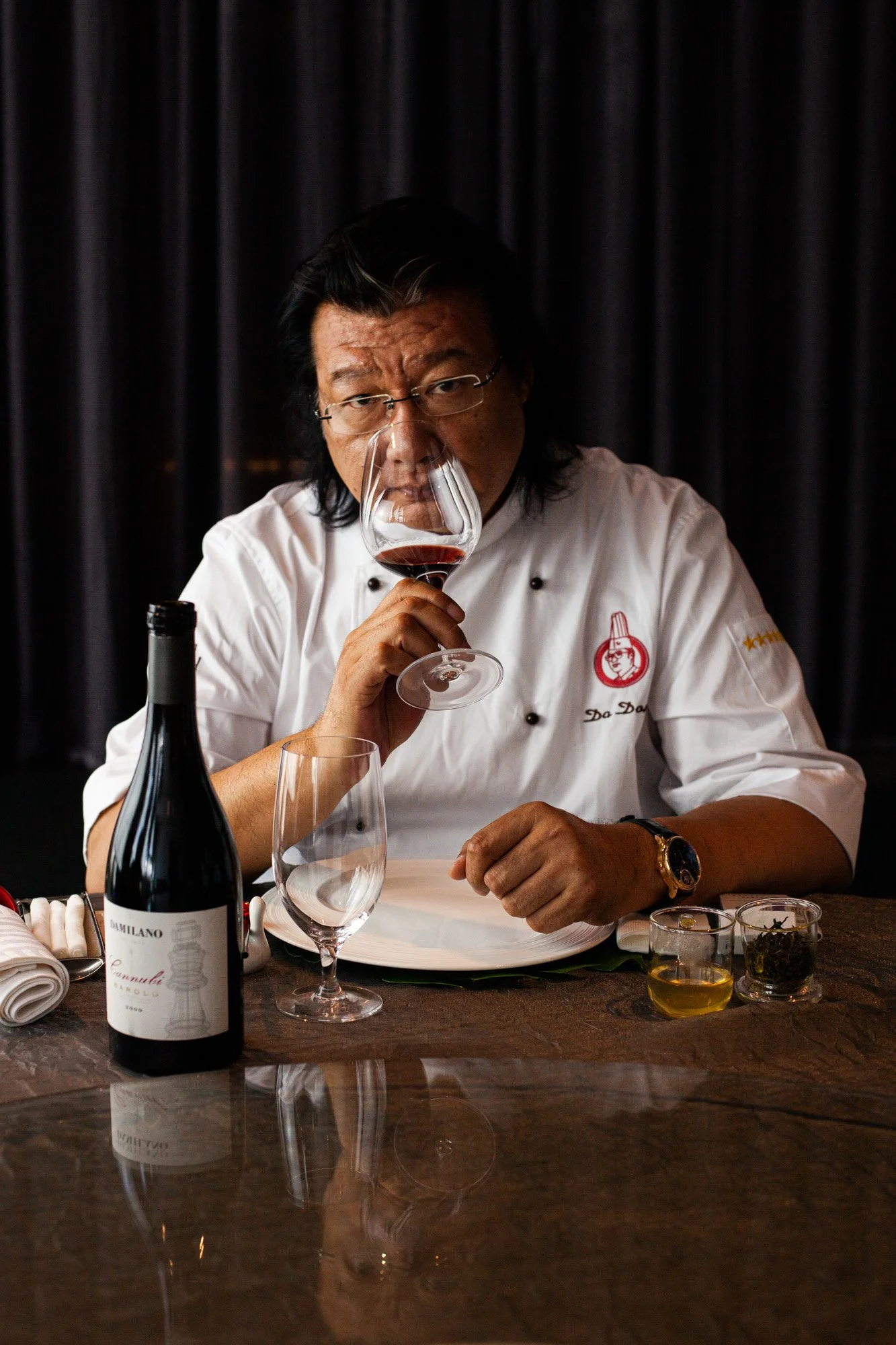 A man in a white chef's coat with glasses and a watch tasting red wine in a dimly lit setting, with a dark curtain in the background and a bottle of wine on the table.