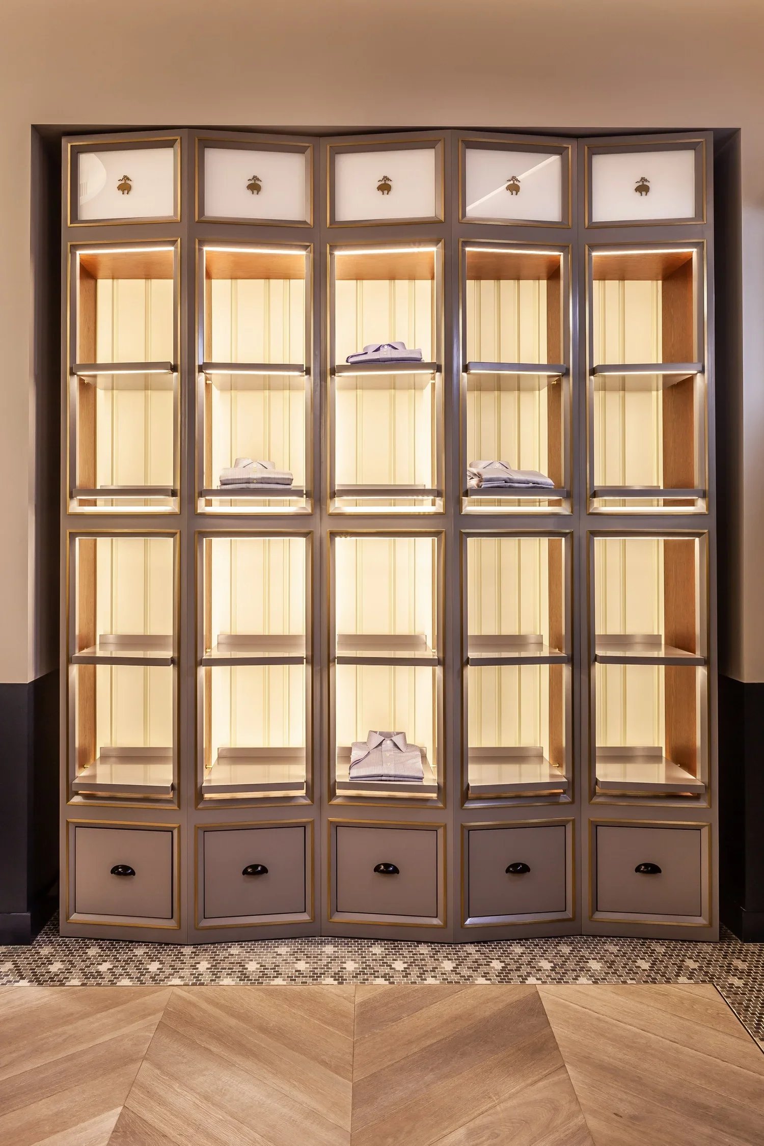 Display shelf with multiple compartments, some containing folded clothing, illuminated with warm lighting, set against a beige wall with a patterned tile floor below.