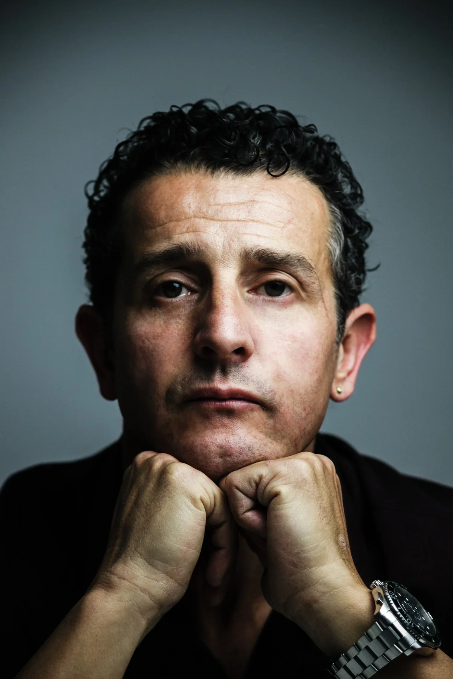 A man with curly hair and earrings, resting his chin on his hands, wearing a silver wristwatch, against a dark background.