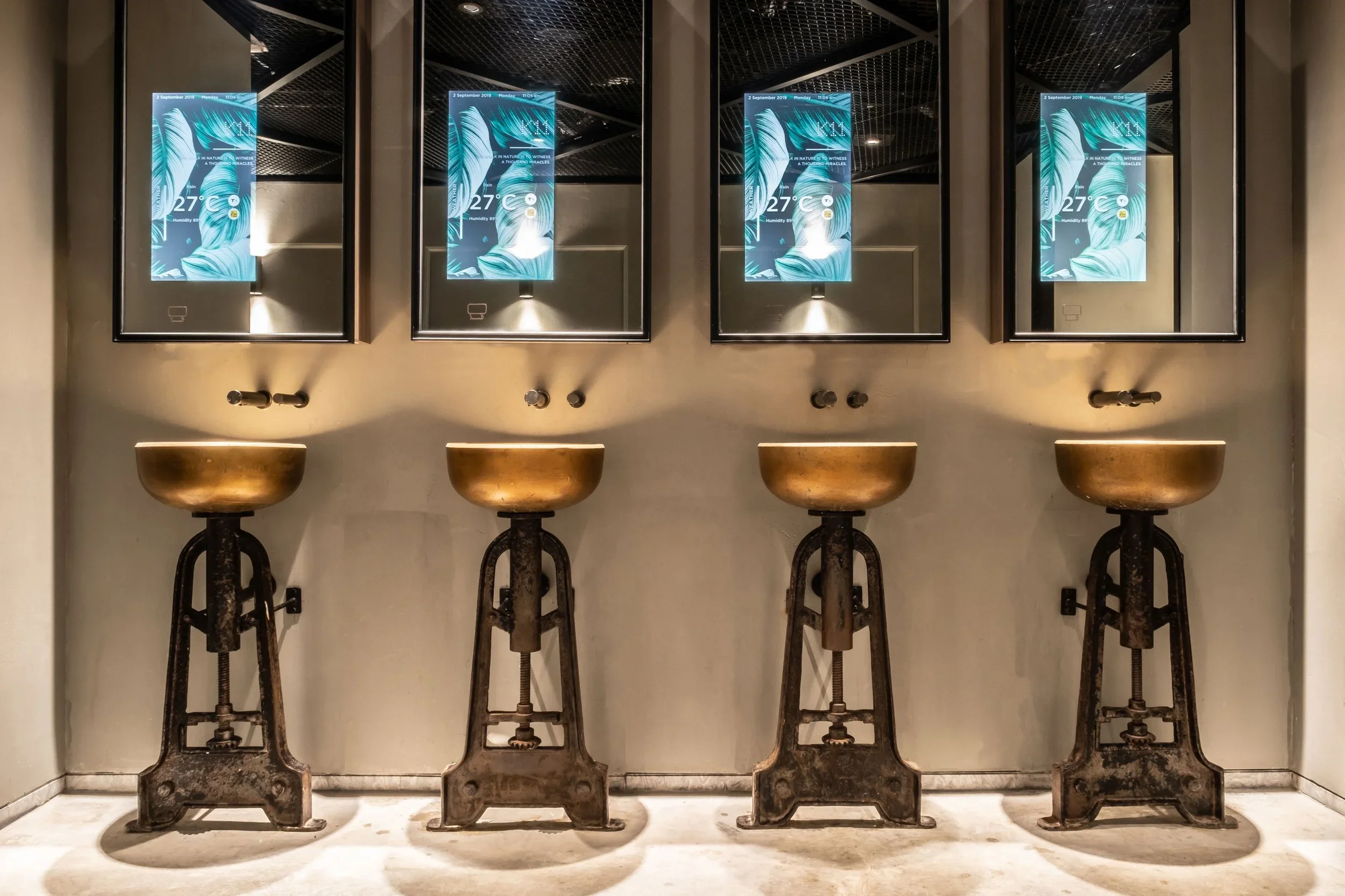 Four vintage sinks with bronze basins and old-fashioned plumbing fixtures mounted on a beige wall, with four framed digital screens displaying a tropical leaf background and weather information above each sink.