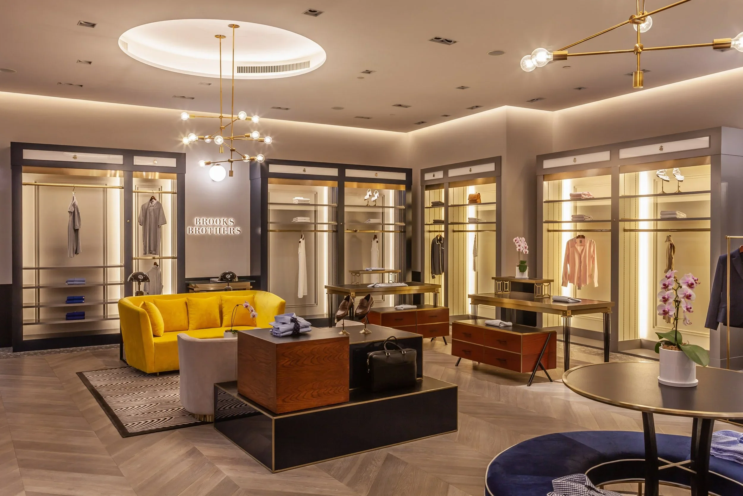 Interior of a high-end clothing boutique with display shelves, hanging garments, a yellow sofa, and modern lighting fixtures.