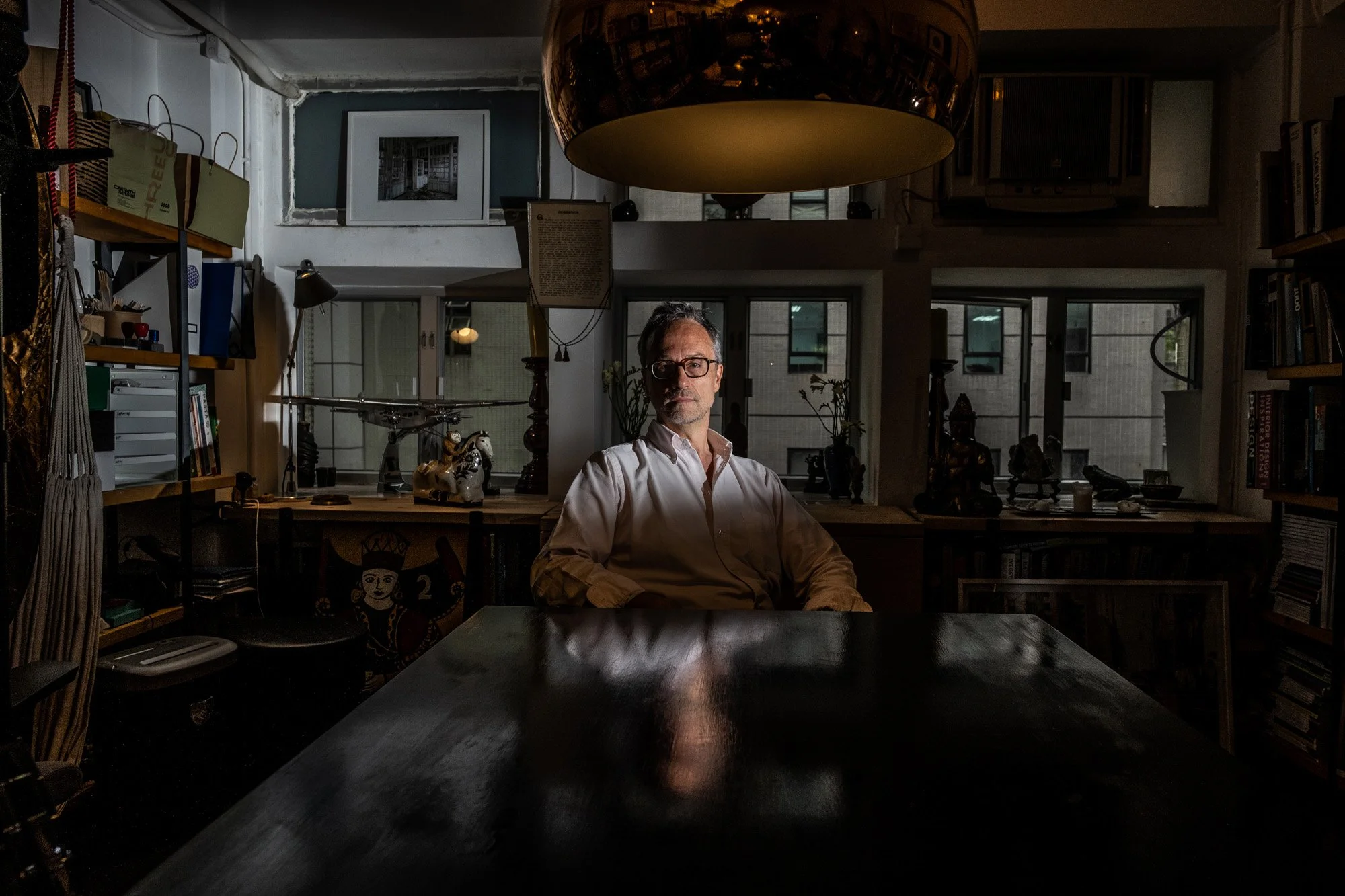 A man with glasses and a white shirt sitting at a dark wooden table in a home office or library, with bookshelves, windows, and decorative items in the background. The room is dimly lit, with natural light coming through the windows.