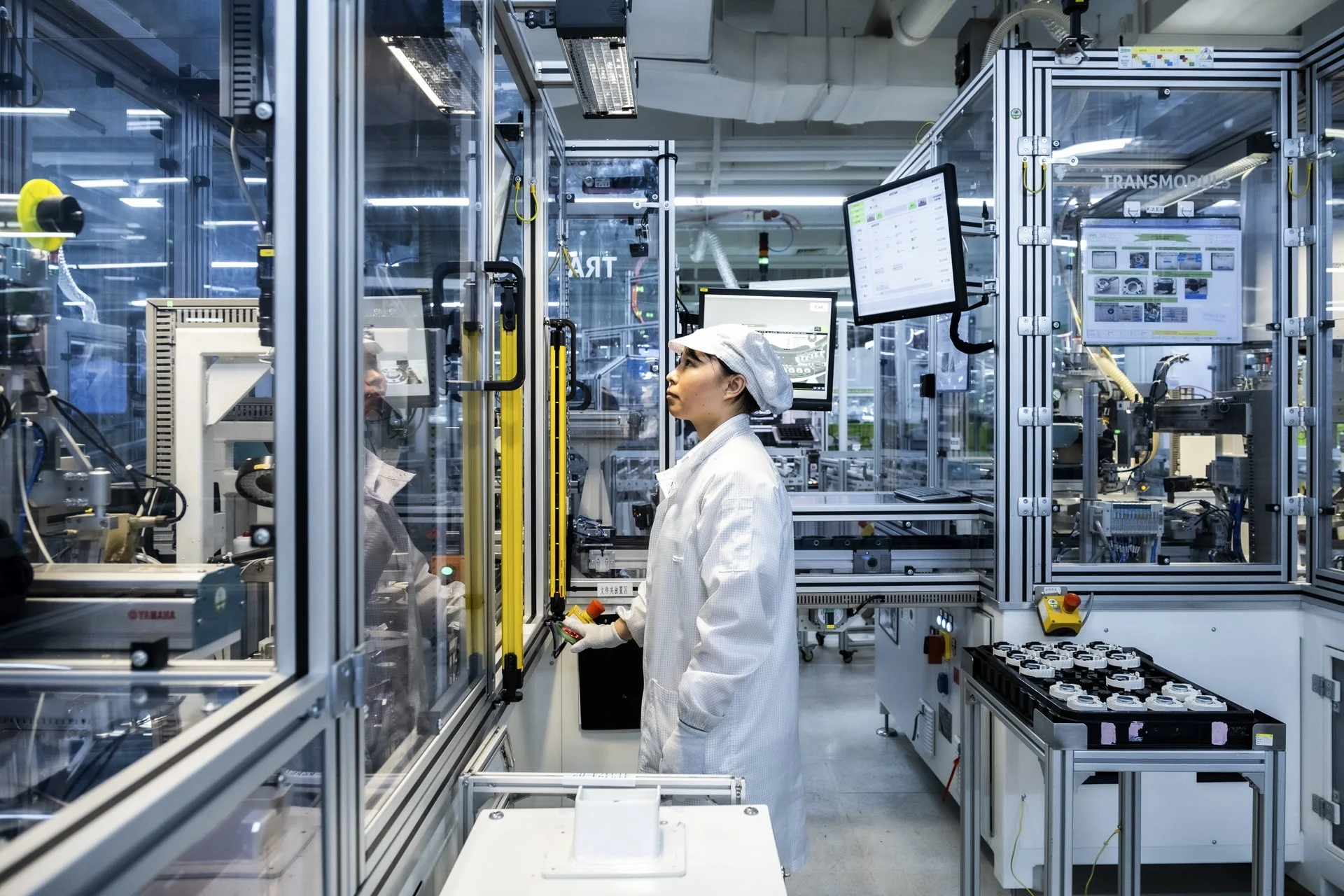 A worker in a white lab coat and hairnet operates machinery inside a high-tech manufacturing facility, with multiple screens displaying data and technical diagrams around them.