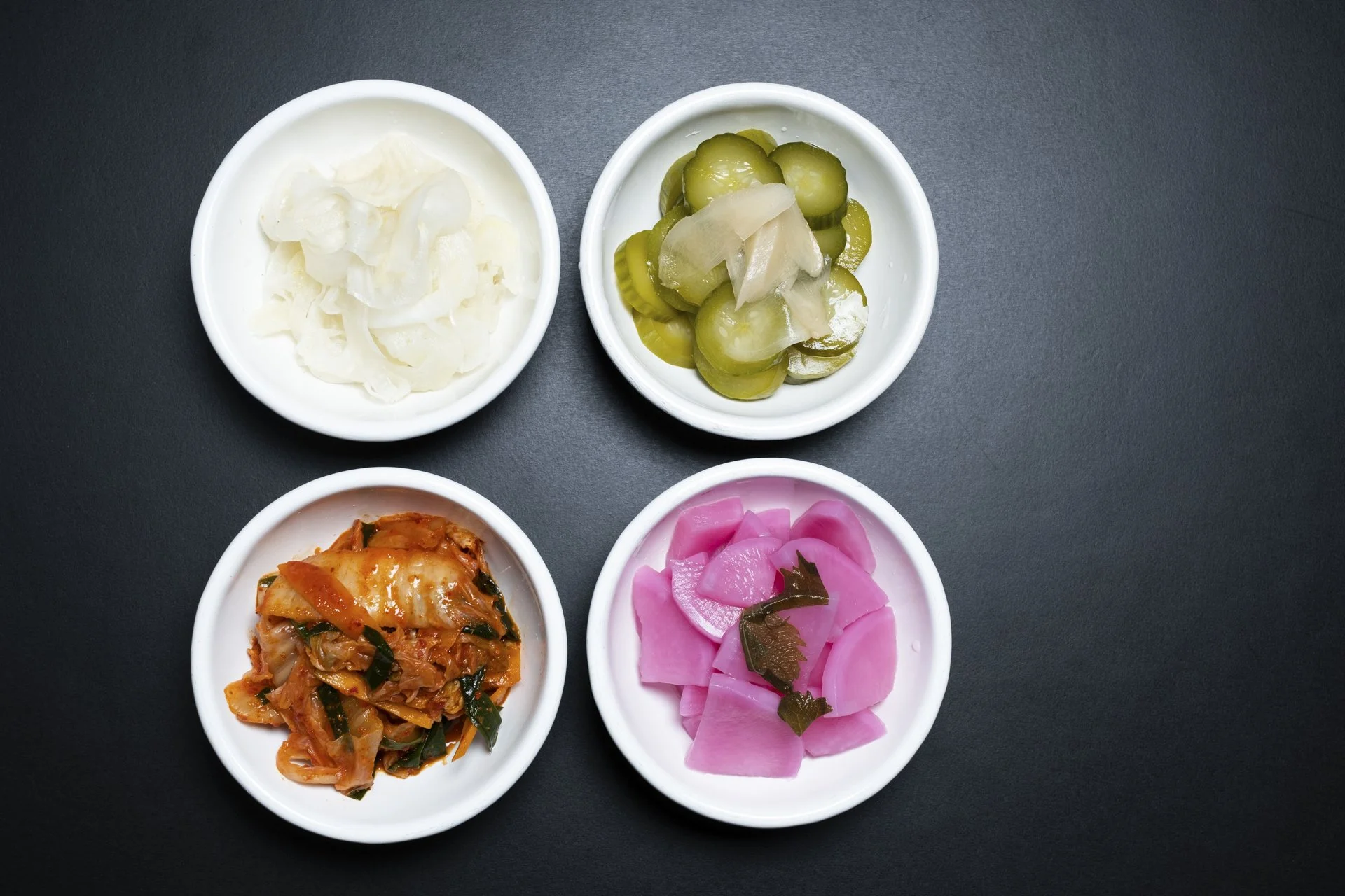 Four small white bowls containing pickled vegetables on a dark surface. Top left bowl has white onions, top right has sliced pickles and daikon radish, bottom left has kimchi, and bottom right has bright pink pickled radish with seaweed garnish.