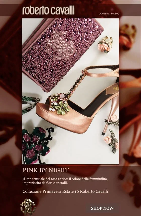 A pink glittery clutch purse, a matching high-heeled shoe with floral embellishments, and floral jewelry pieces arranged on a white background, with a promotional text for Roberto Cavalli's Spring 2023 collection.
