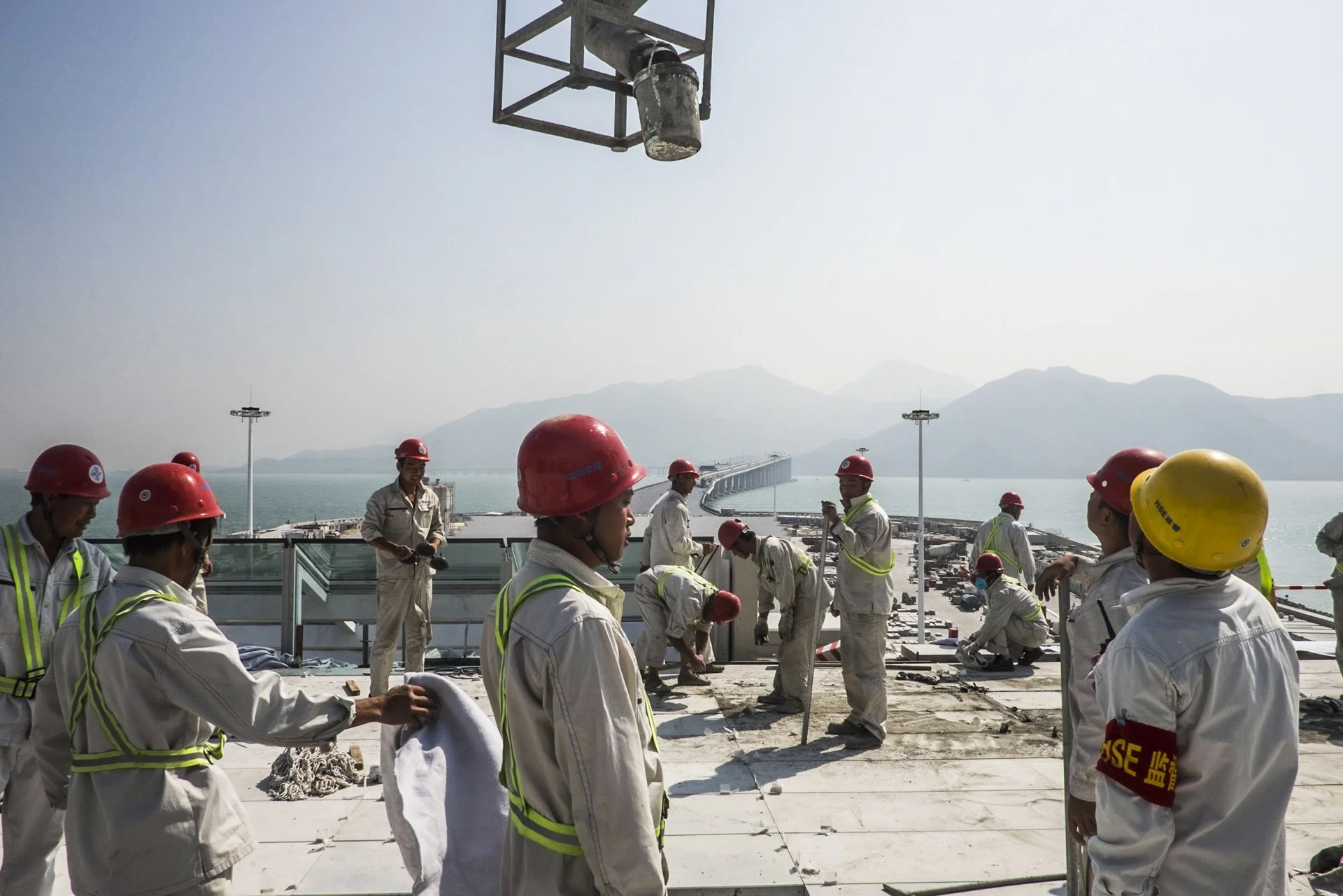 Construction workers wearing safety helmets and vests working on a building site near a body of water with mountains in the background.