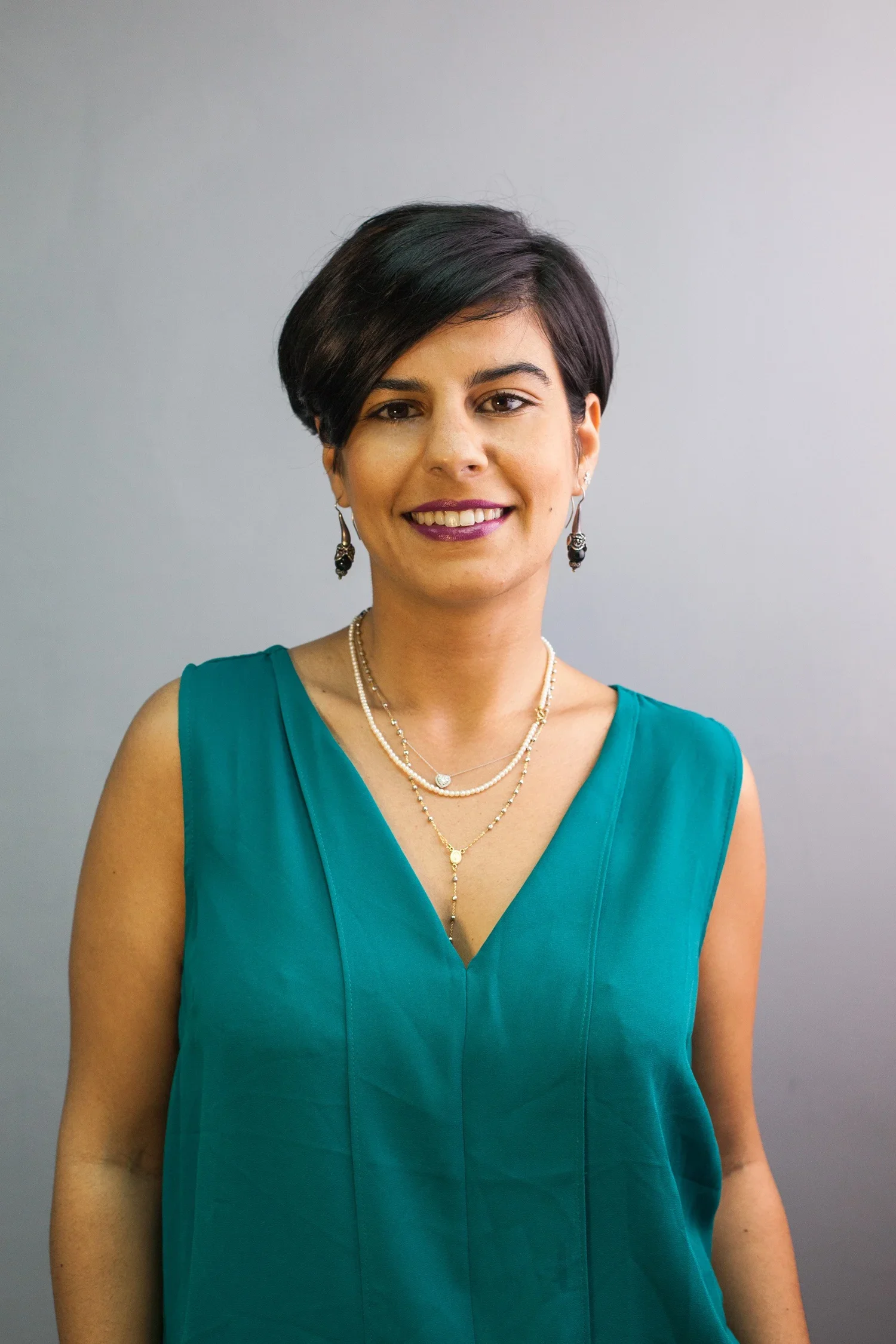 A woman wearing a teal sleeveless dress and layered necklaces, smiling in front of a gray background.