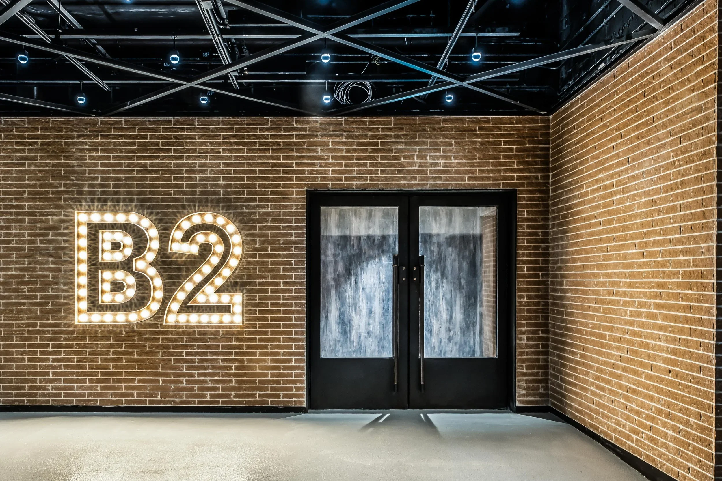 Interior view of a building with a brick wall, illuminated sign with the text 'B2', and a black door, modern ceiling with exposed wiring and lighting.