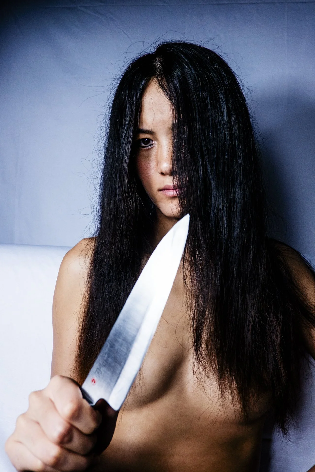 A woman with long black hair, partly covering her face, holding a large kitchen knife pointed towards her face, against a plain background.