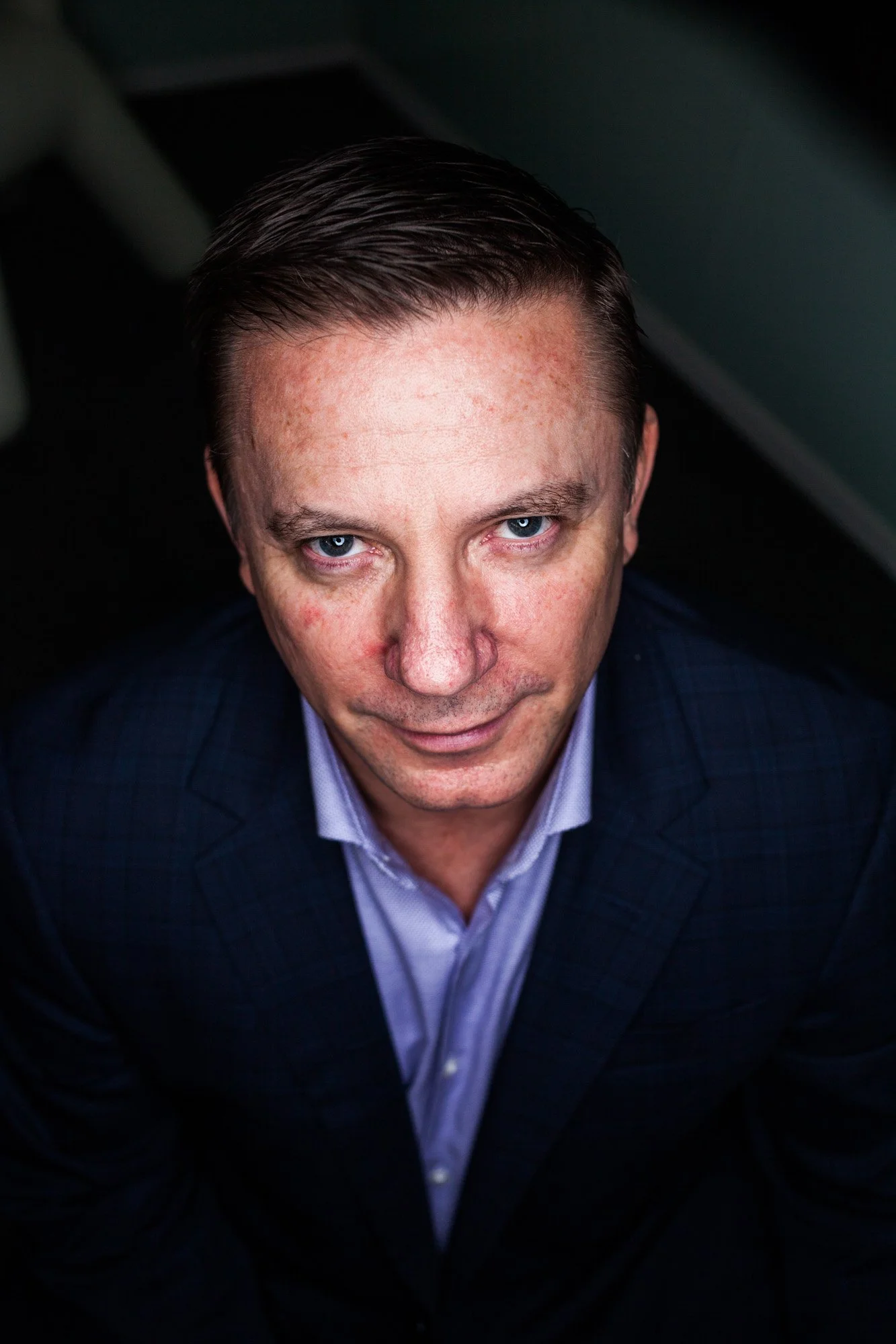 Close-up portrait of a man with blue eyes, short dark hair, wearing a navy blue suit and light purple shirt, looking directly at the camera, against a dark background.
