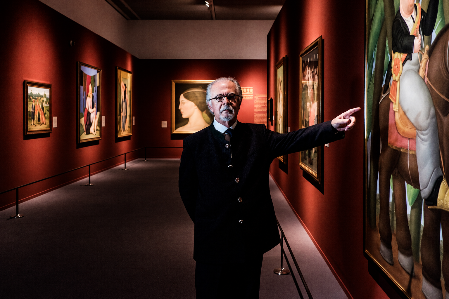 An older man with glasses and a gray beard, wearing a black suit, is standing in an art gallery with red walls. He is pointing at a painting on the wall to his right, surrounded by other framed artworks.