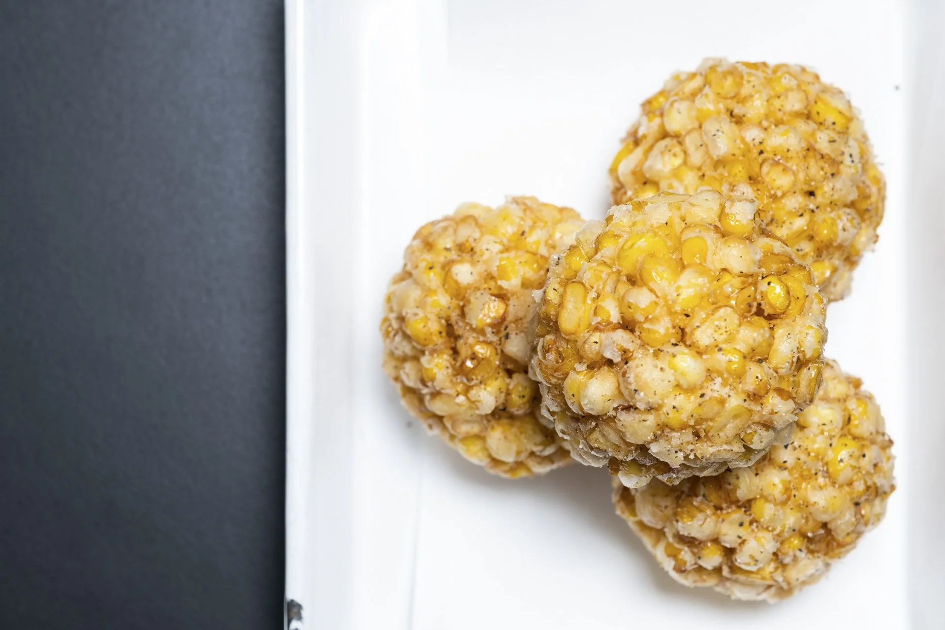 Close-up of round rice cereal treats with corn and black pepper on a white rectangular plate against a dark background.