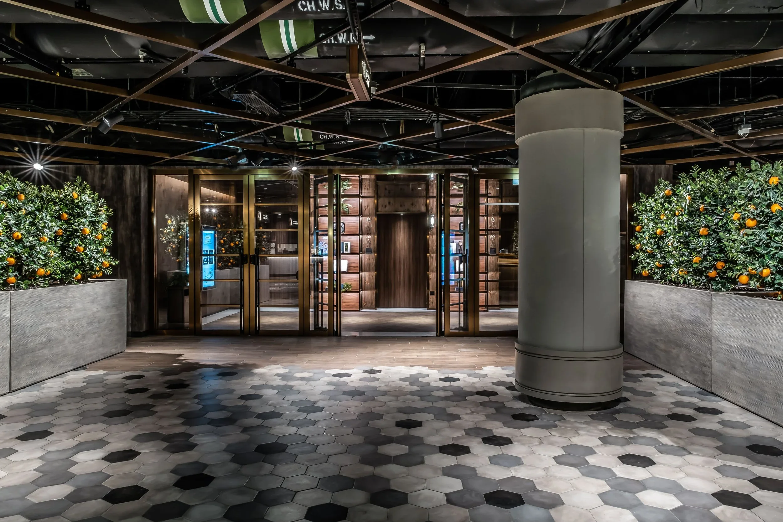 Interior view of a modern restaurant or café with a black ceiling, wooden framework, and spotlights. Two large planters with orange trees flank a set of glass doors with wooden framing, leading to another area. The floor has a hexagonal tile pattern 