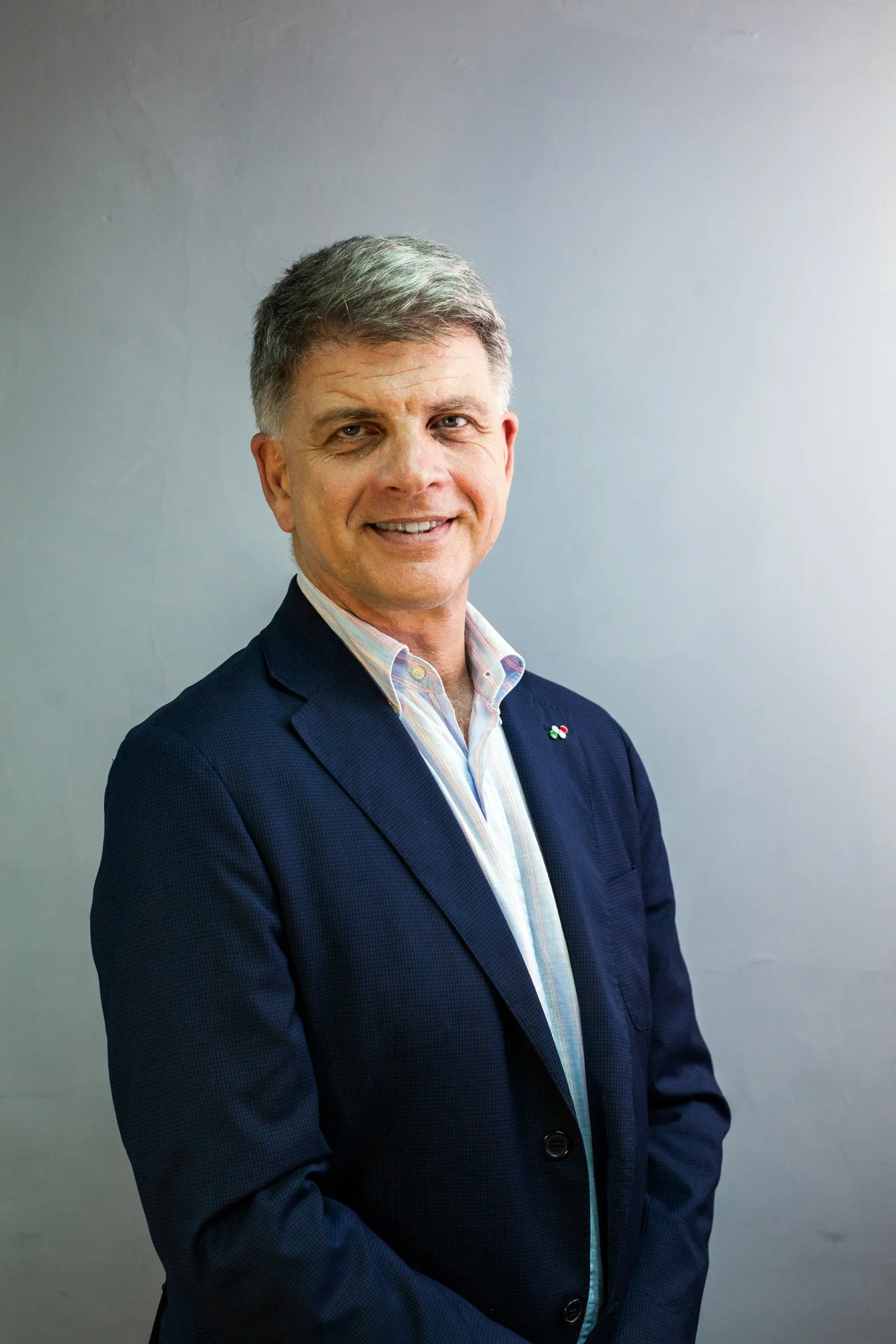A middle-aged man with gray hair, wearing a navy blue blazer and a light-colored striped shirt, standing against a plain gray wall, smiling slightly with arms crossed.