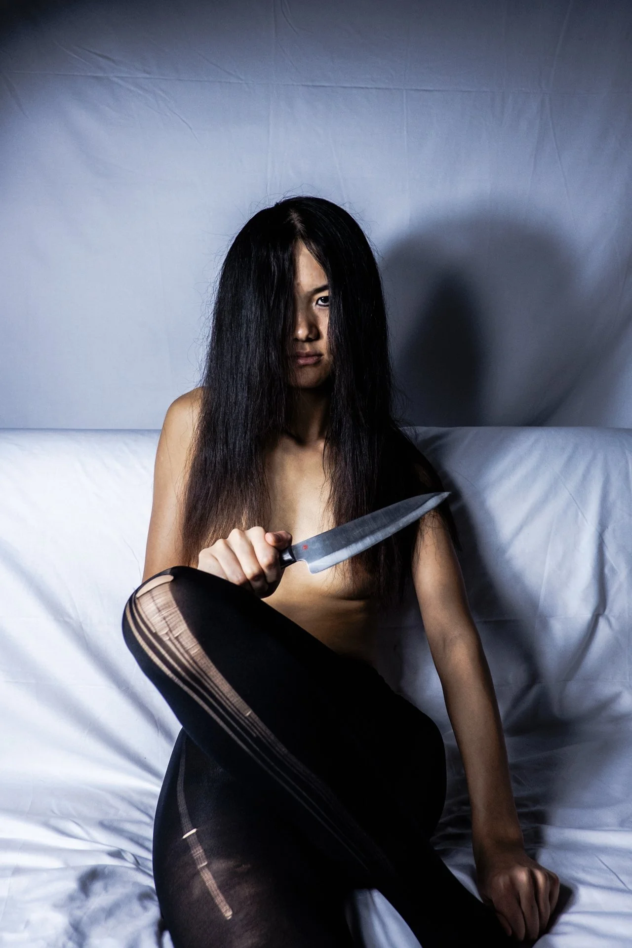 A woman with long black hair, partially covering her face, holding a large kitchen knife. She is sitting on a bed with white sheets, wearing black ripped stockings, in a dimly lit room with a gray backdrop.
