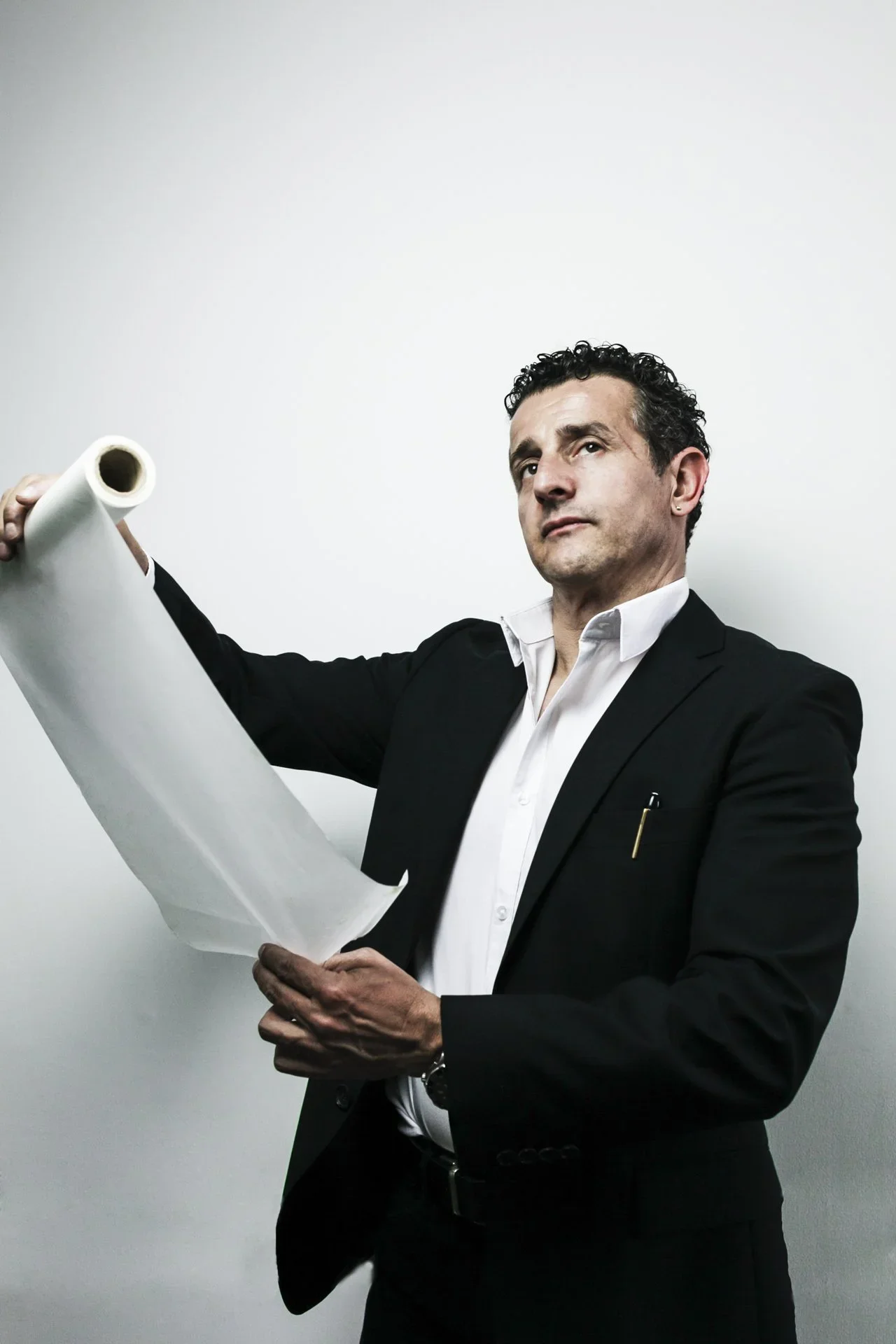A man in a black suit and white shirt holding a large rolled-up blueprint or architectural plan, standing against a plain white background.