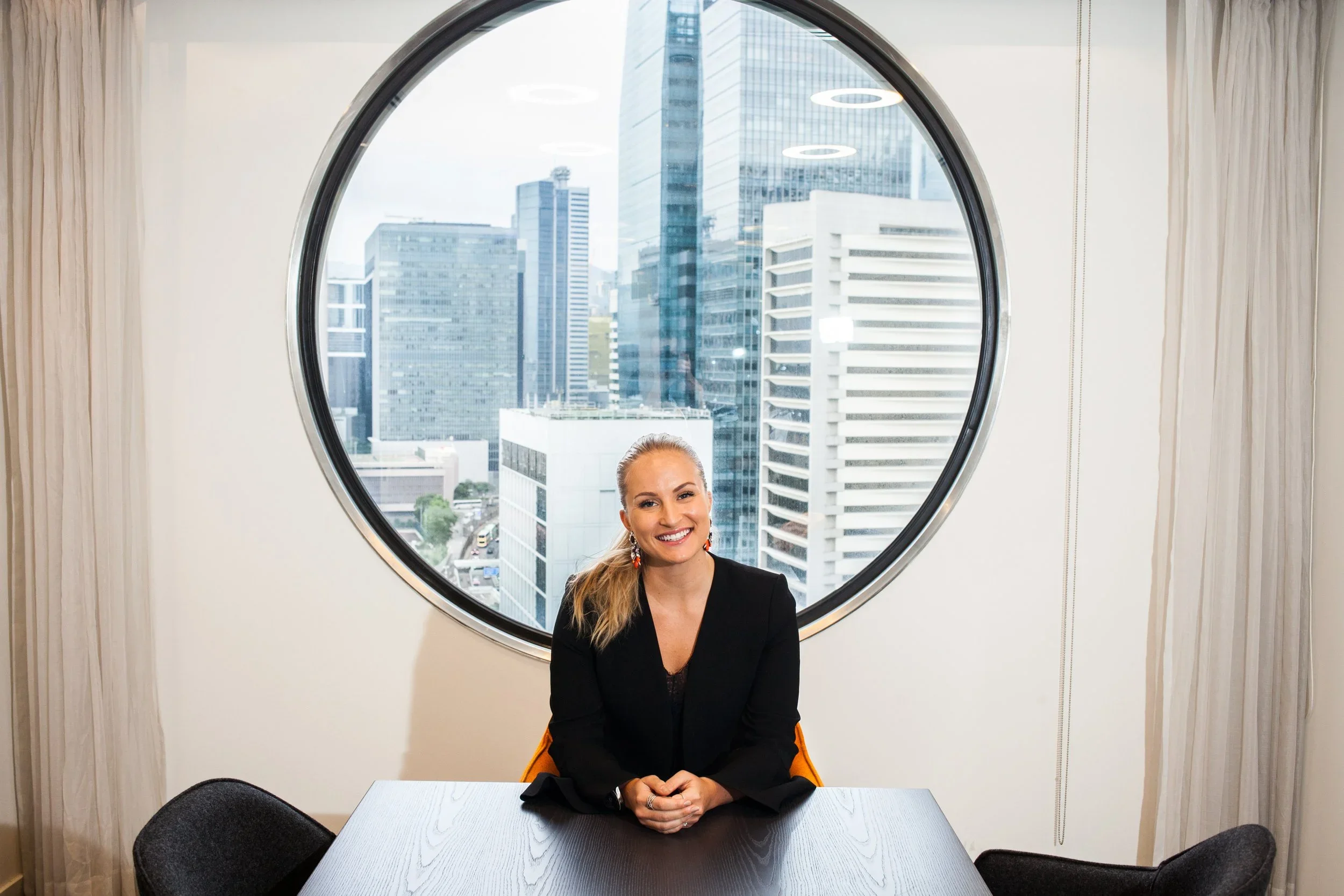 A woman with blonde hair, smiling, sitting at a table in a room with a large oval window showing a cityscape with tall buildings.