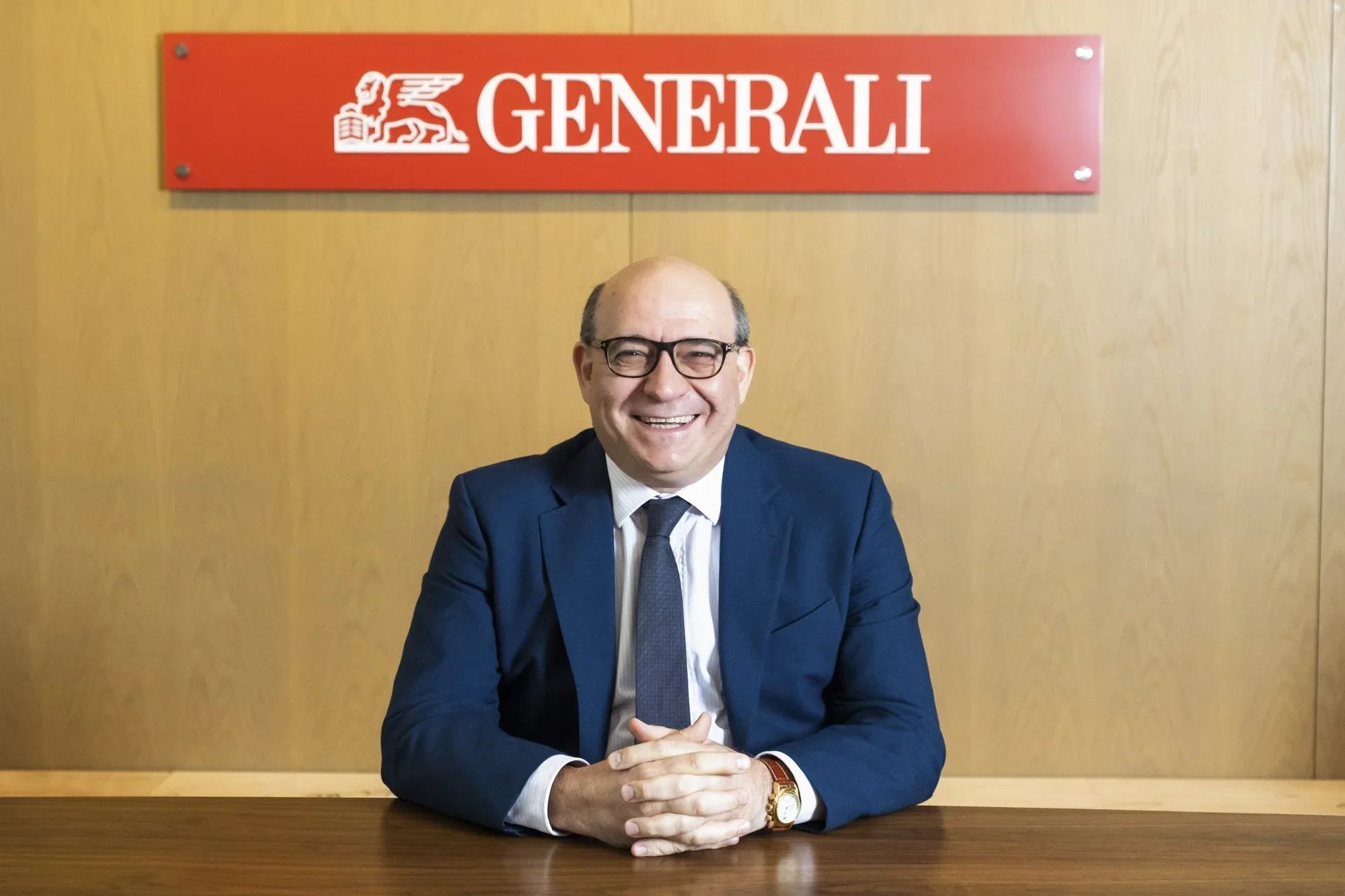 A smiling man in a suit and glasses sitting at a wooden table with hands clasped in front of him. Behind him is a red sign with the logo and name of Generali insurance company on a beige wall.