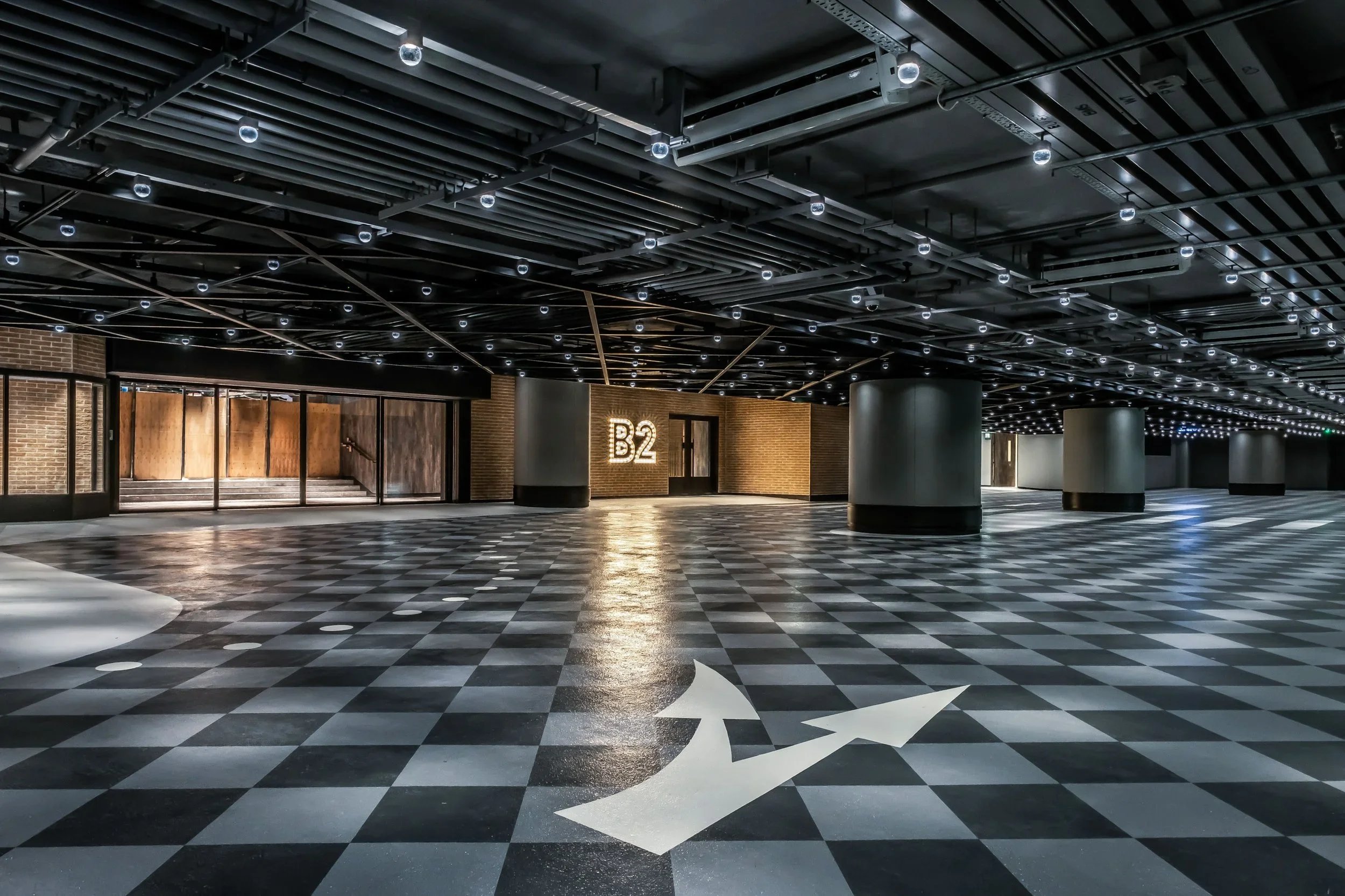 Empty indoor space with black and white checkered floor, ceiling with exposed pipes and small circular lights, and a lit sign reading "B2" on the brick wall in the background.