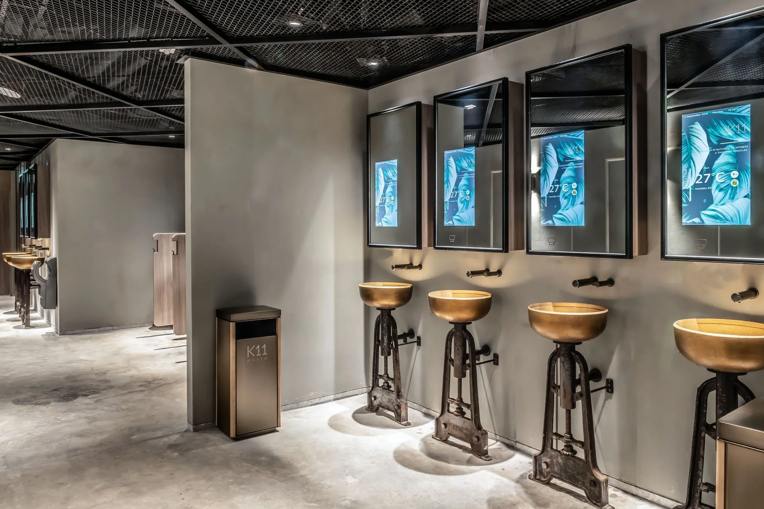 Interior of a modern building with digital screens displaying weather info and four rustic sinks with wash basins and fixtures