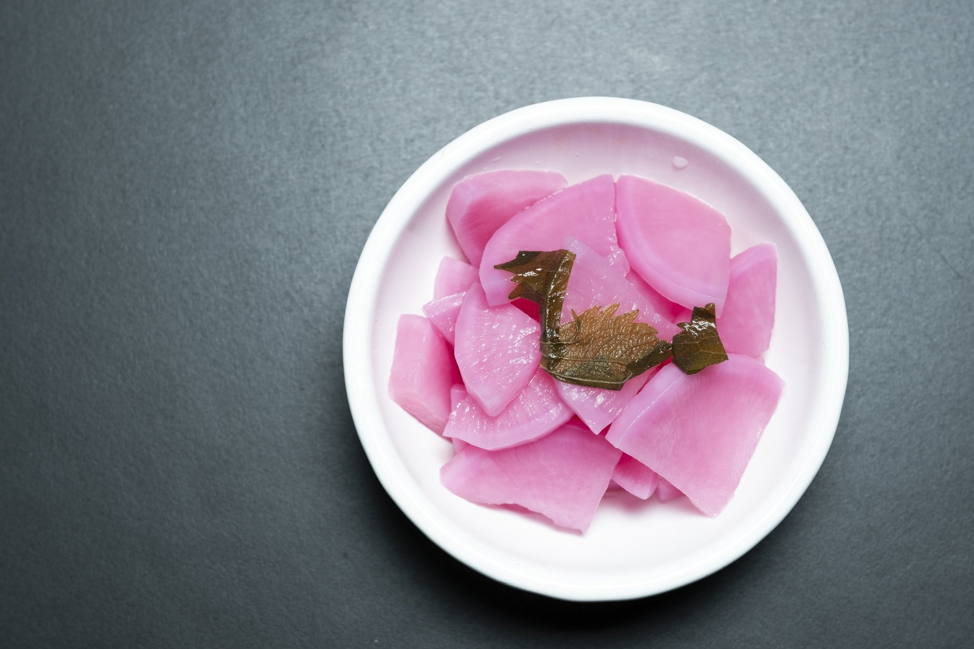A white bowl containing pickled pink radish slices garnished with dark green seaweed on a dark surface.