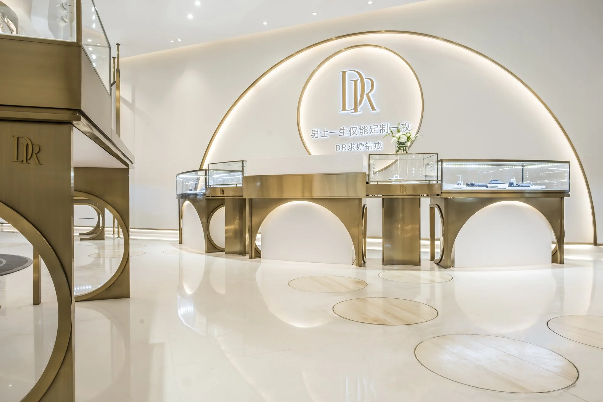 Luxurious jewelry display area with glass cases on golden stands, a white wall with a backlit logo 'DR,' and elegant lighting and decor.