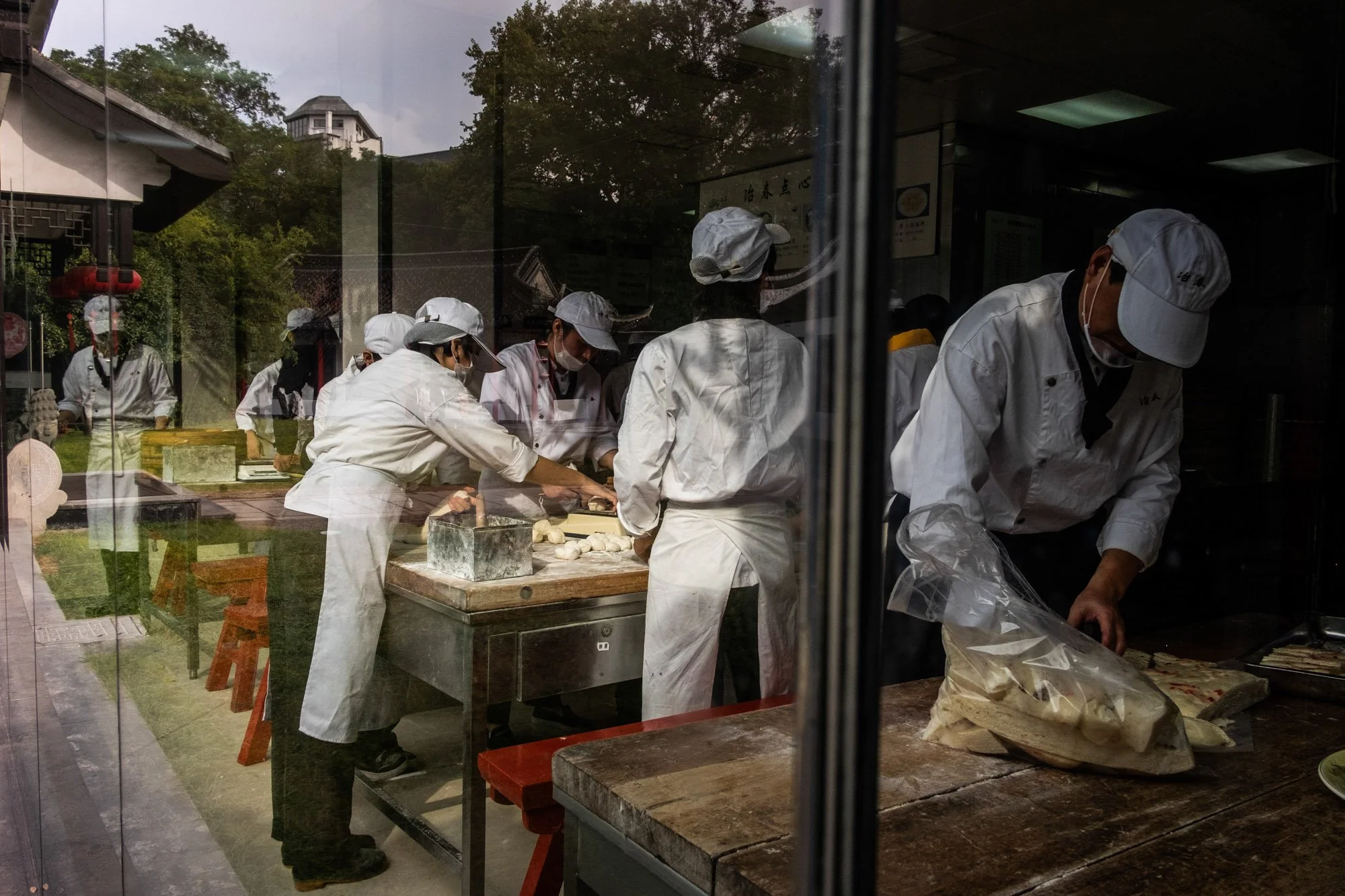 Chefs in white uniforms and hats preparing and cooking food inside a restaurant kitchen, seen through a window.