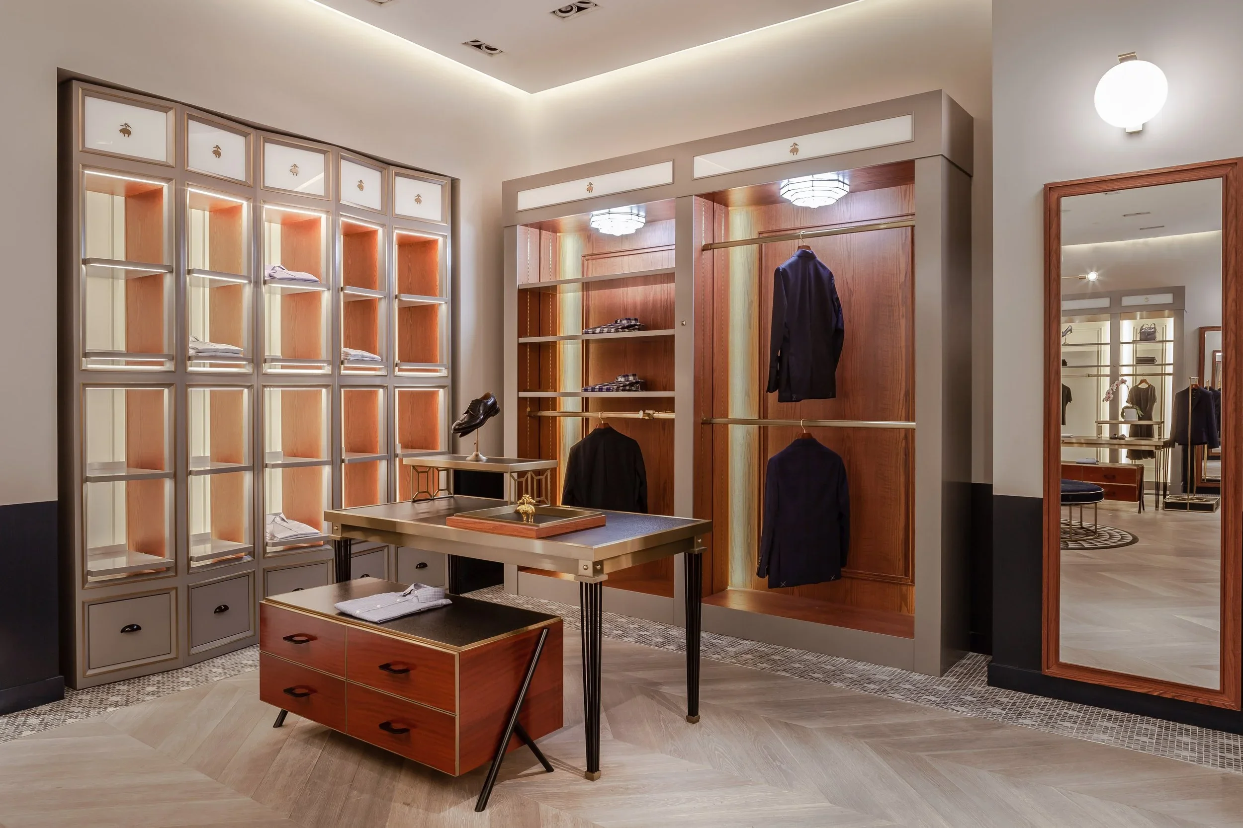 A retail clothing store with wooden shelves, racks of suits, and a large mirror. The store has bright lighting with modern fixtures and a neutral color palette.