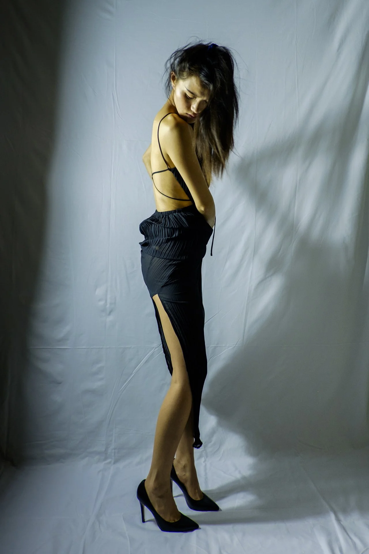 A woman with long dark hair wearing black high heels, black sheer skirt with thigh slit, and a black strappy top, standing against a plain light-colored backdrop.