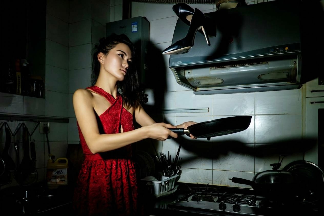 A woman in a red dress cooking in a kitchen, holding a frying pan over a stove.