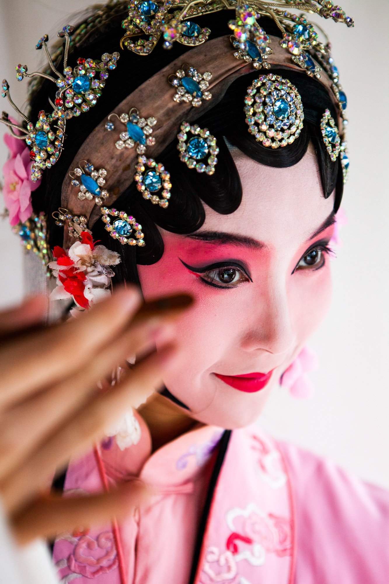 Close-up of a woman dressed in traditional Chinese opera costume with pink makeup, ornate headpiece adorned with colorful gemstones and flowers, and elegant eye makeup.