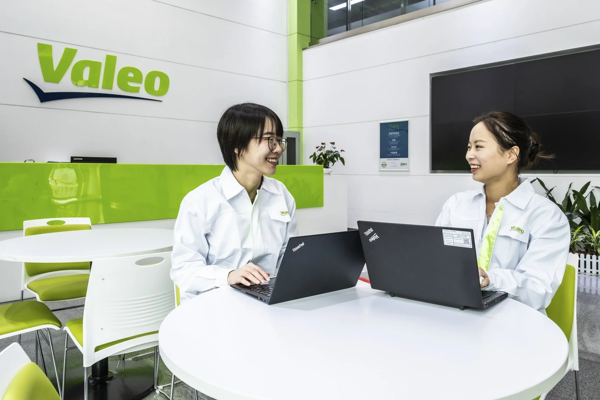 Two women wearing white uniforms with the Valeo logo, working on laptops and smiling while seated at a white round table in a modern office with white and green decor.