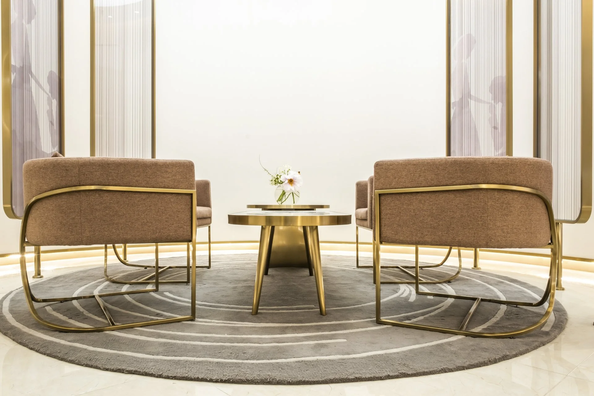 Modern waiting area with two pink upholstered armchairs with gold metal frames, a round gold table with a floral arrangement, a circular rug, and a white backdrop with vertical gold accents.