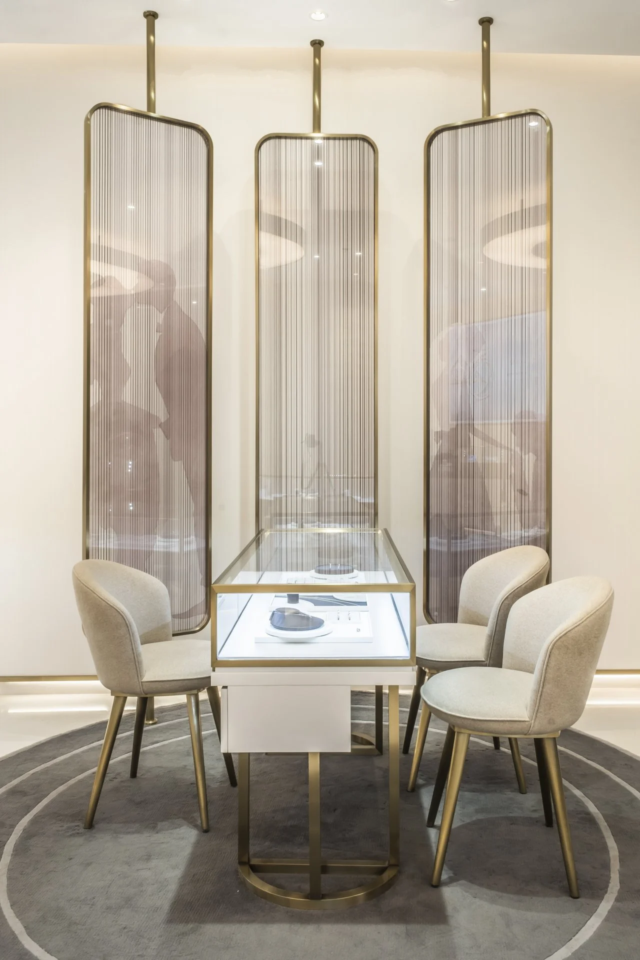 Elegant seating area with four beige upholstered chairs surrounding a white table with glass display case, set on a circular gray rug, with decorative panels and soft lighting in the background.