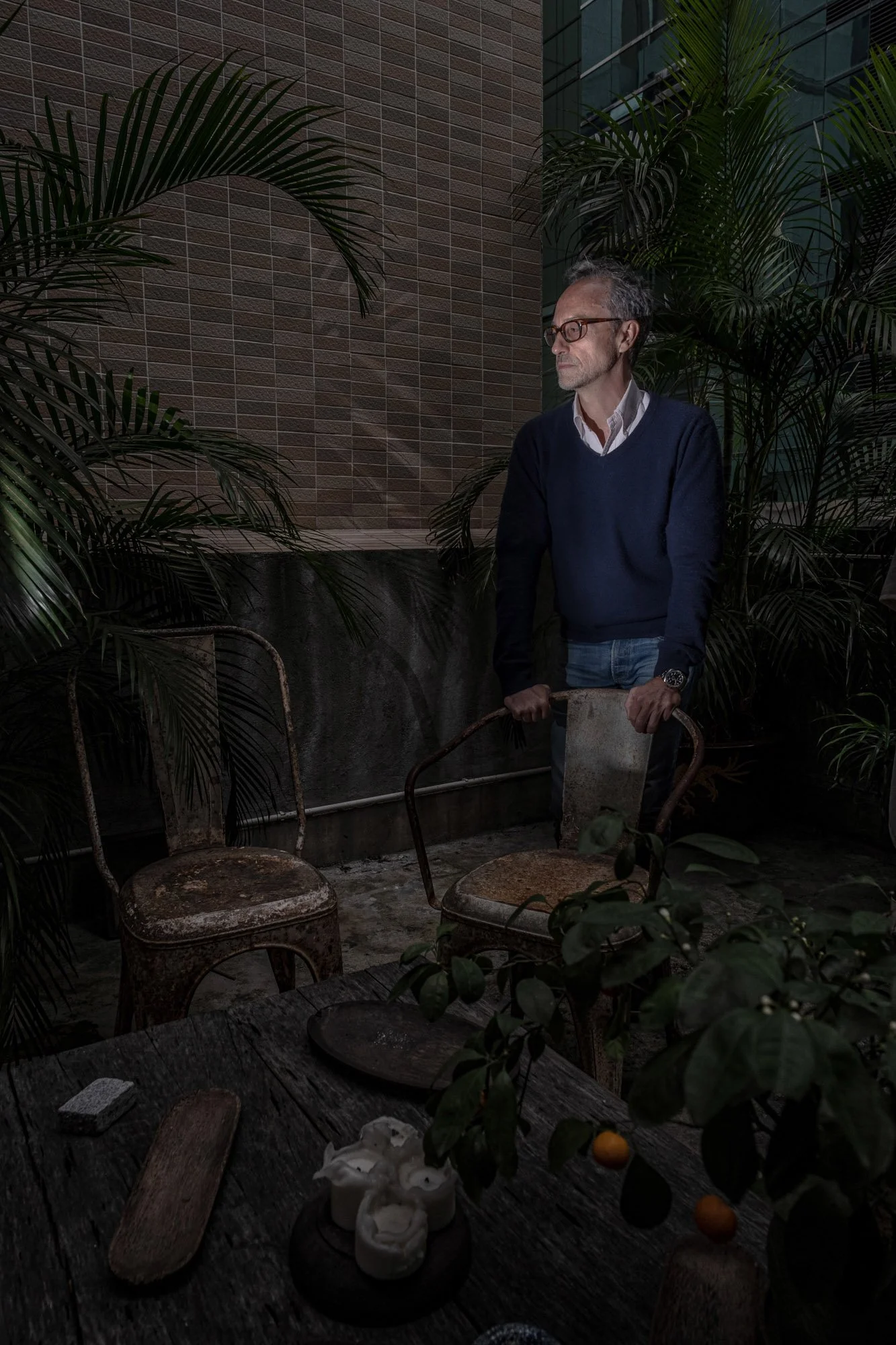 A man wearing glasses, a white shirt, and a blue sweater stands outdoors among plants with a brick and glass building in the background. He is holding a rusty metal chair.