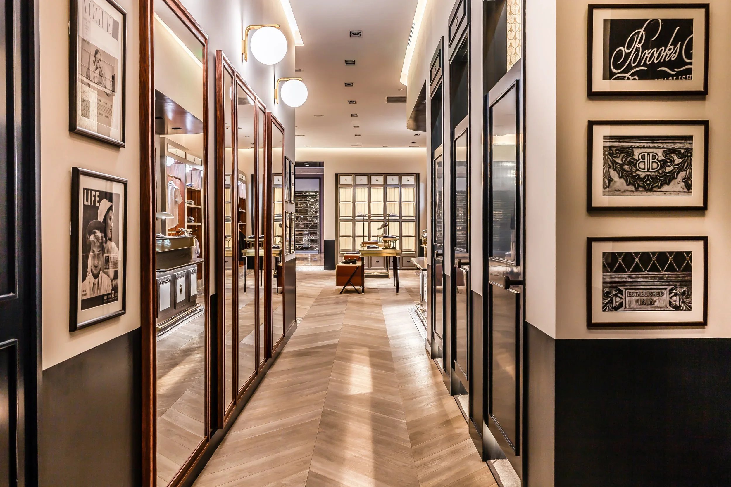 A modern interior with framed vintage photographs on walls, wooden panels, large round wall sconces, and a room with desks and a window at the back.