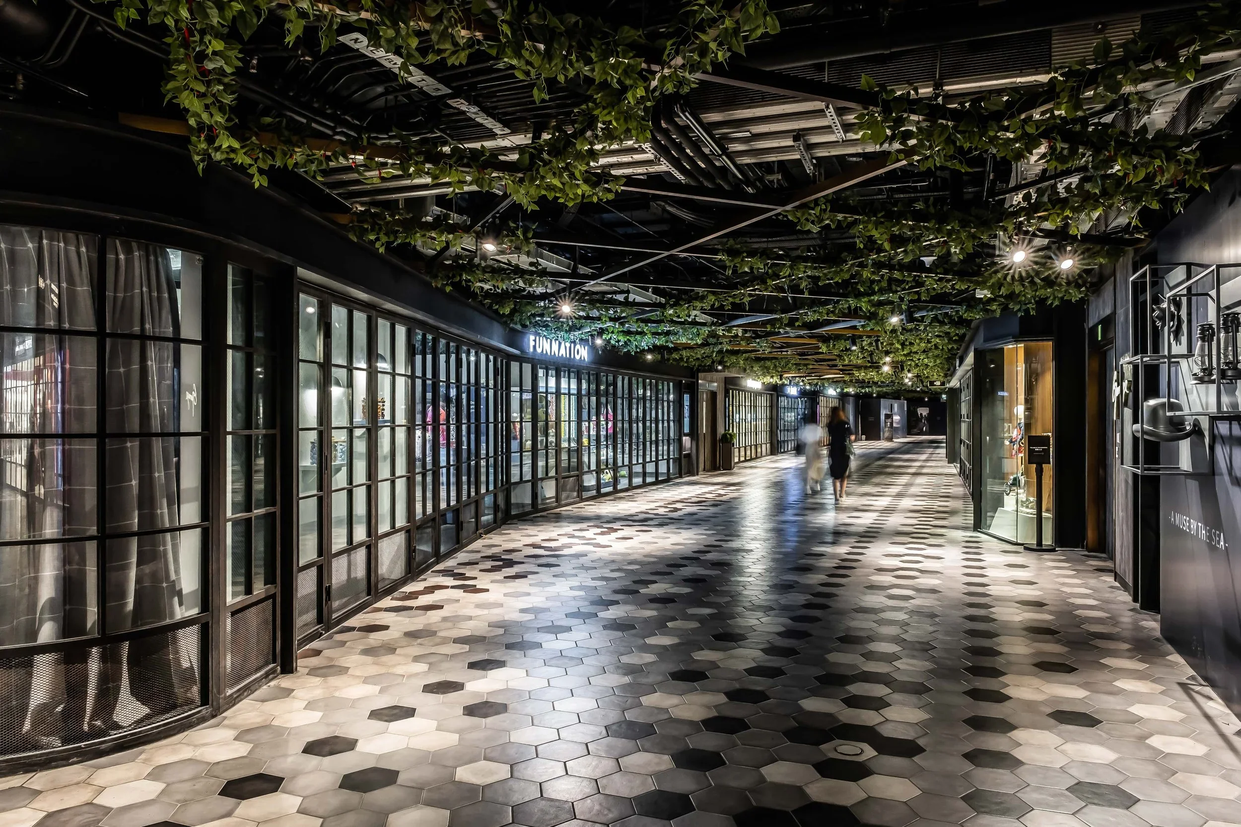 Empty shopping mall corridor with glass storefronts, hanging greenery on the ceiling, and a black, white, and gray tile floor.